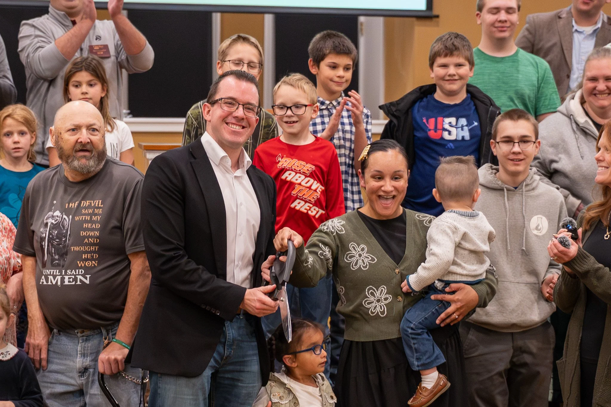 A group of children and adults gathered in a classroom, with some smiling and clapping. A woman in the center holds a young child, and a man in a suit is handing her a pair of scissors, indicating a celebration or ceremony.