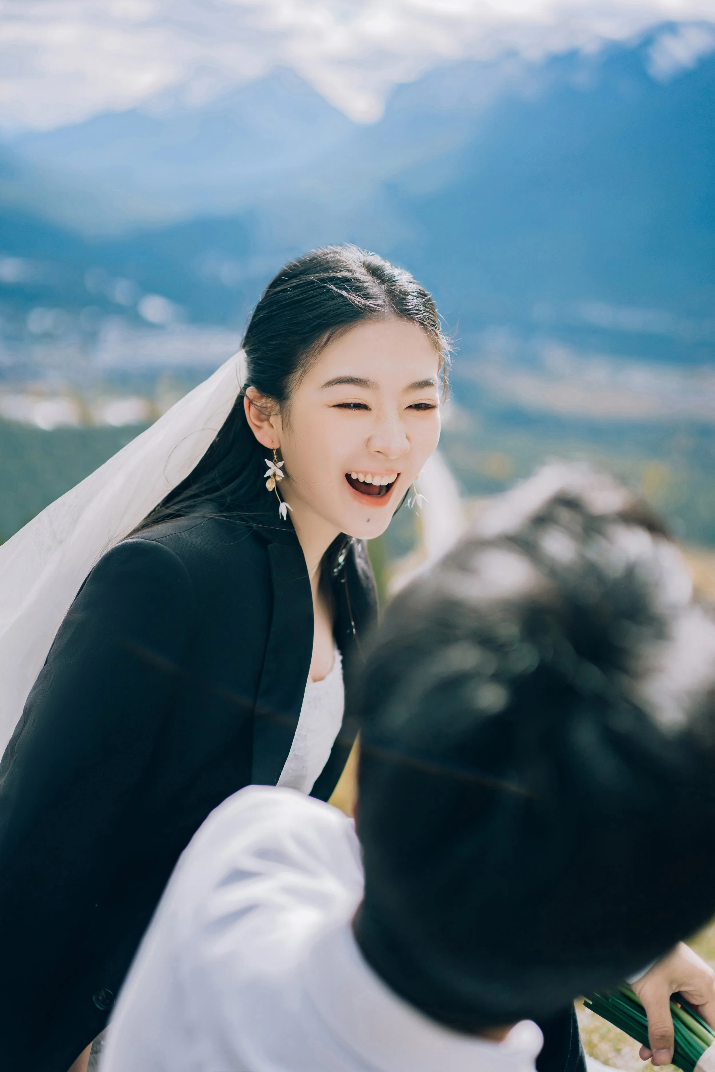 A woman dressed in wedding attire, smiling and laughing outdoors with mountains in the background.