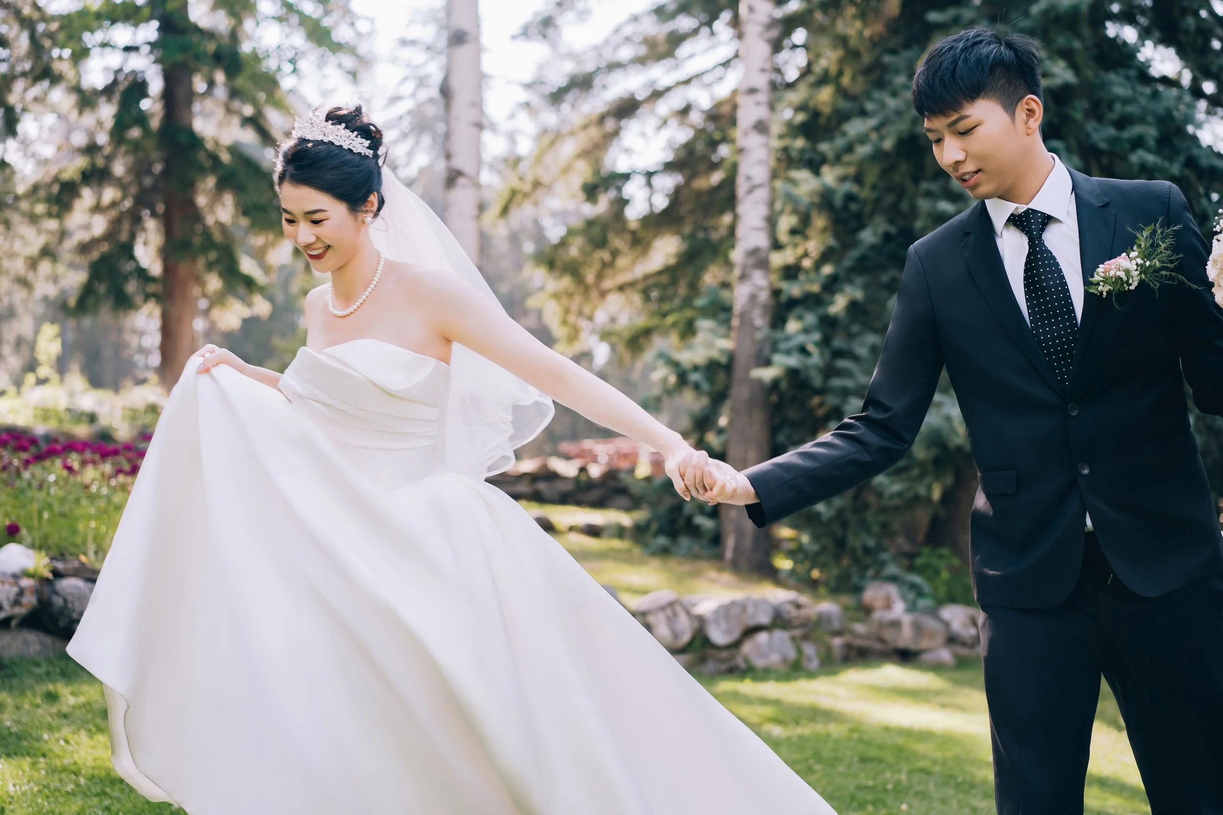A bride in a wedding dress and groom in a suit holding hands outdoors in a park-shaped setting with trees and rocks, smiling and happy.