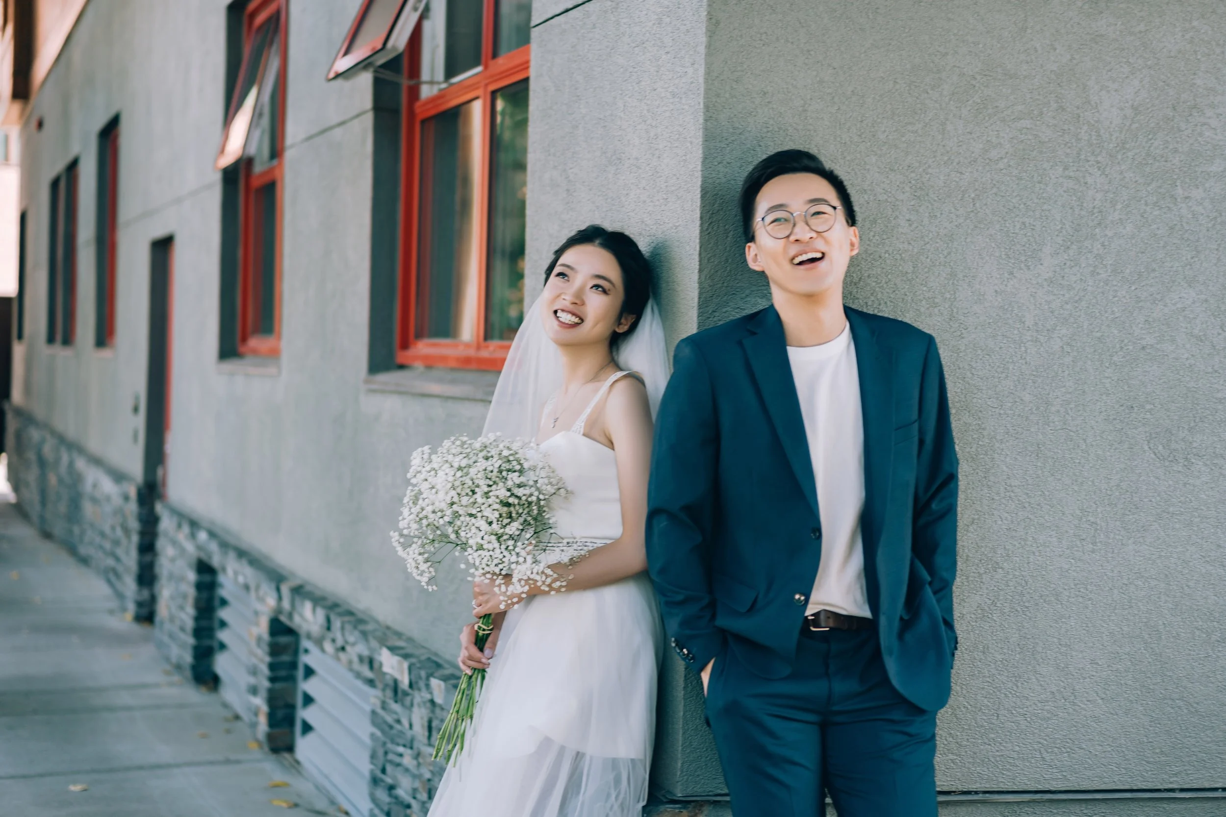 A smiling bride in a white wedding dress holding a bouquet of white flowers, leaning against a building with a groom in a blue suit and glasses, both enjoying a happy moment outdoors.