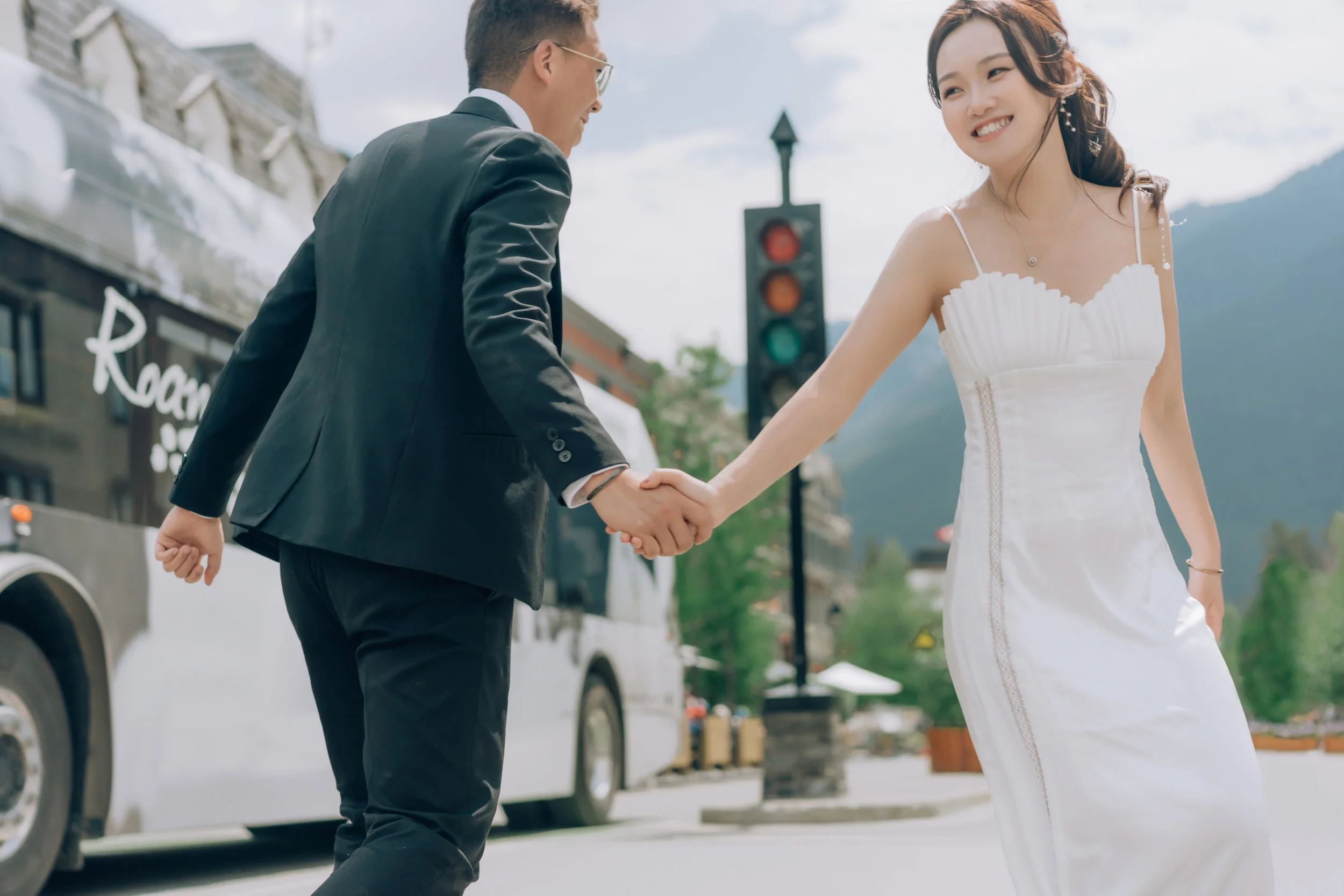 A man and woman are holding hands and smiling, standing outdoors near a bus and a traffic light with mountains in the background. The woman is wearing a white dress, and the man is dressed in a black suit.