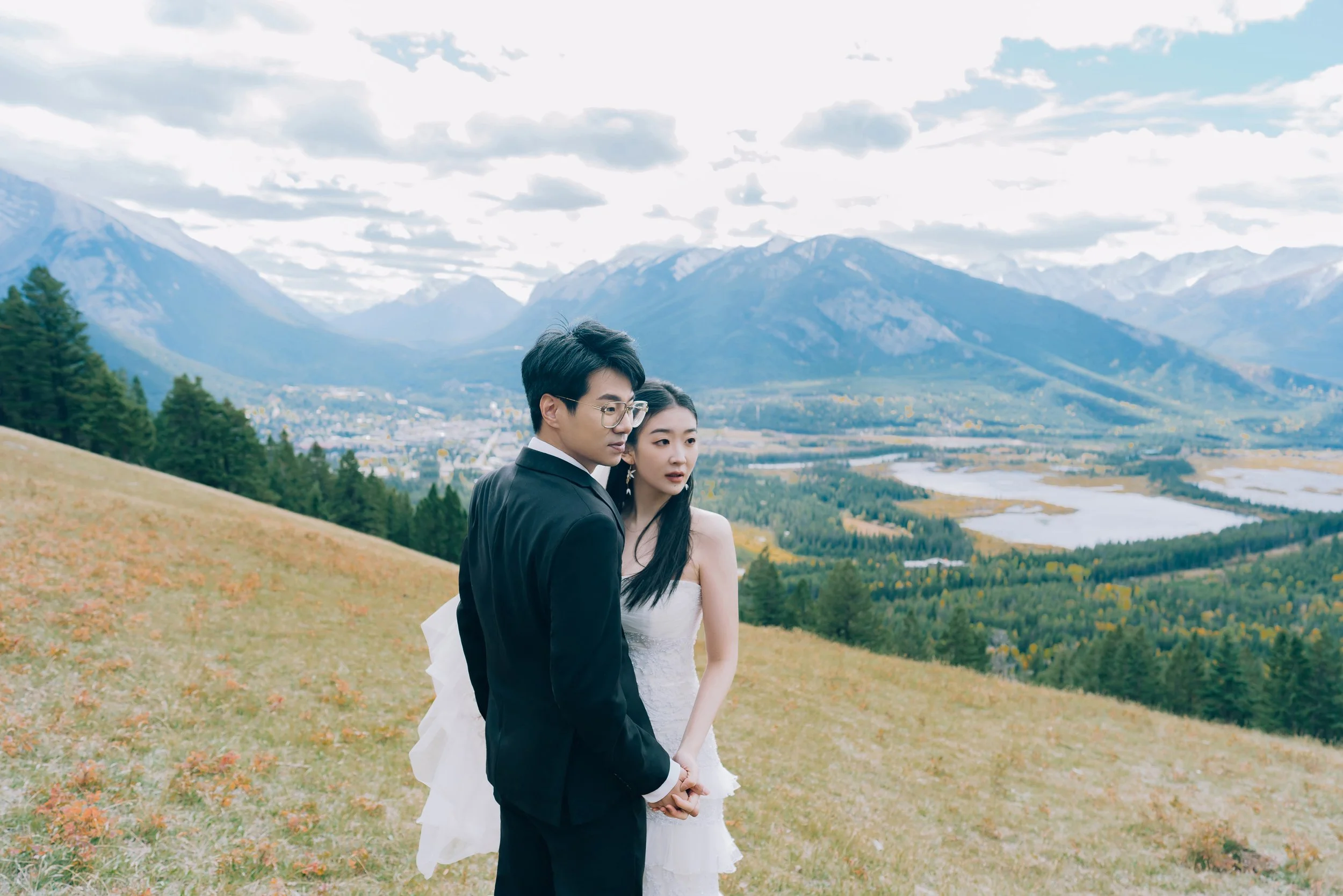 A couple dressed in wedding attire stands on a grassy hillside with mountains, trees, and a river in the background.