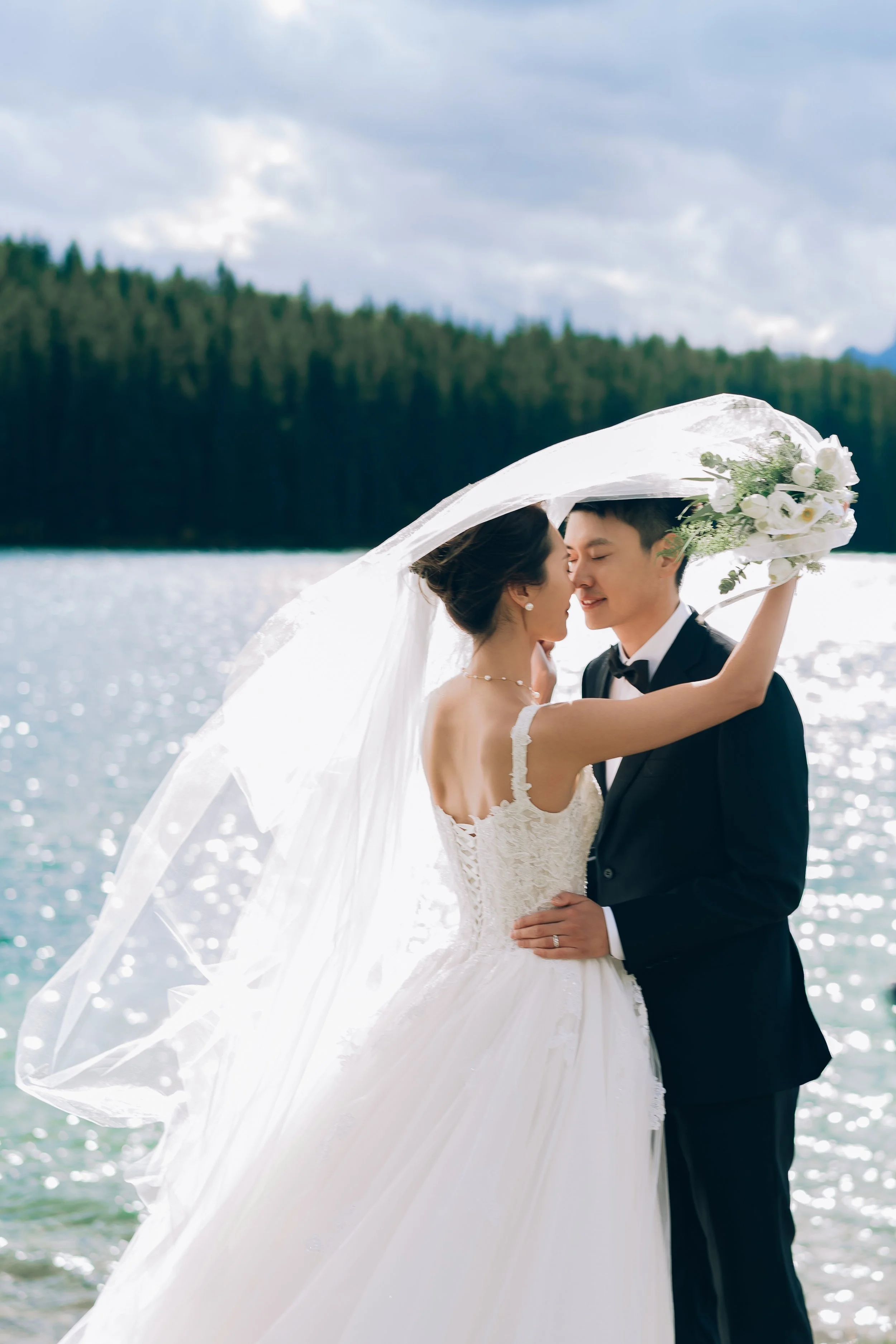 A bride and groom in wedding attire embrace by a lake with trees in the background, under an umbrella.
