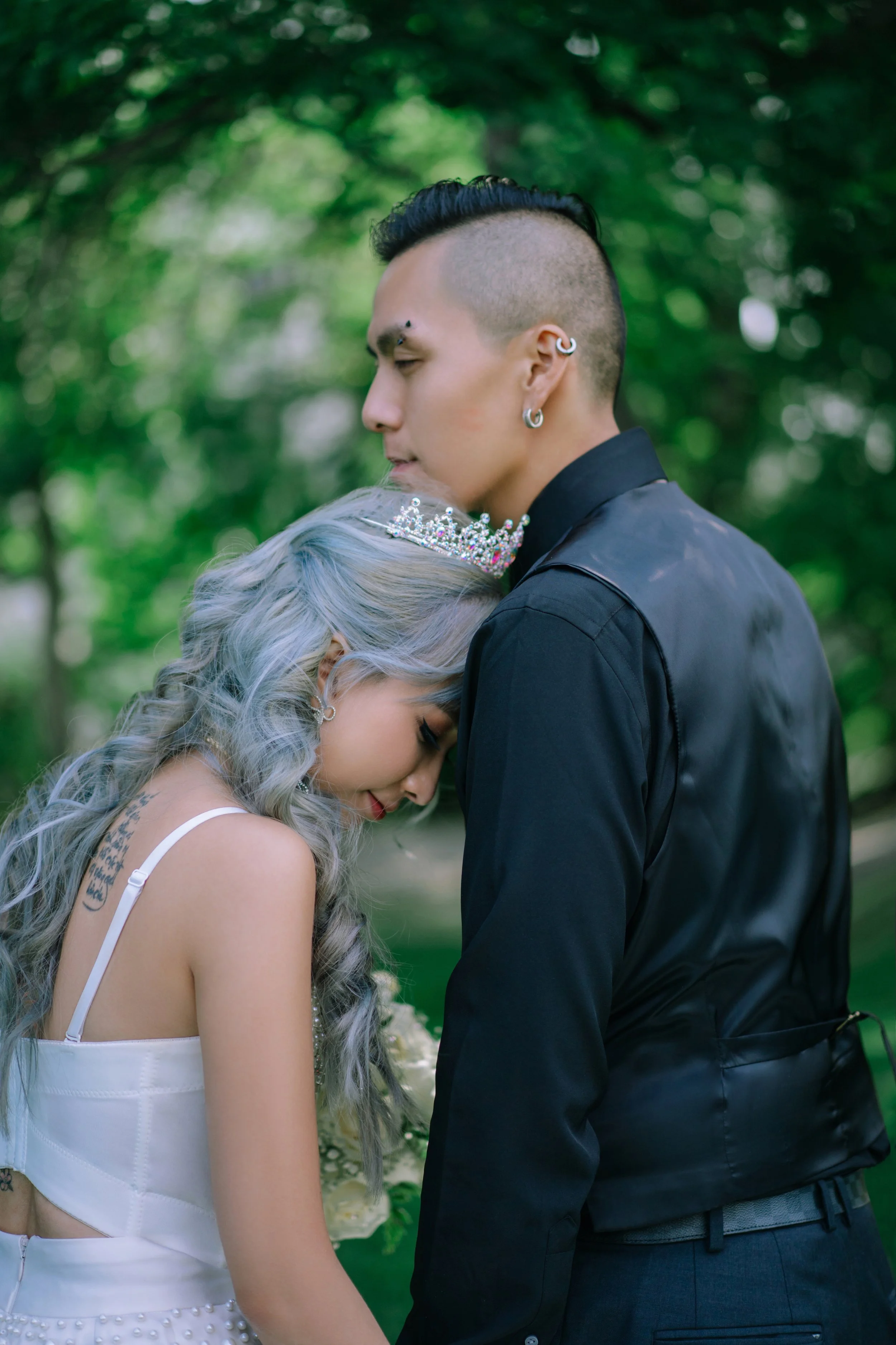 A couple with unique hairstyles in a close embrace outdoors, the woman wearing a tiara and the man in a black shirt with earrings, surrounded by green trees.