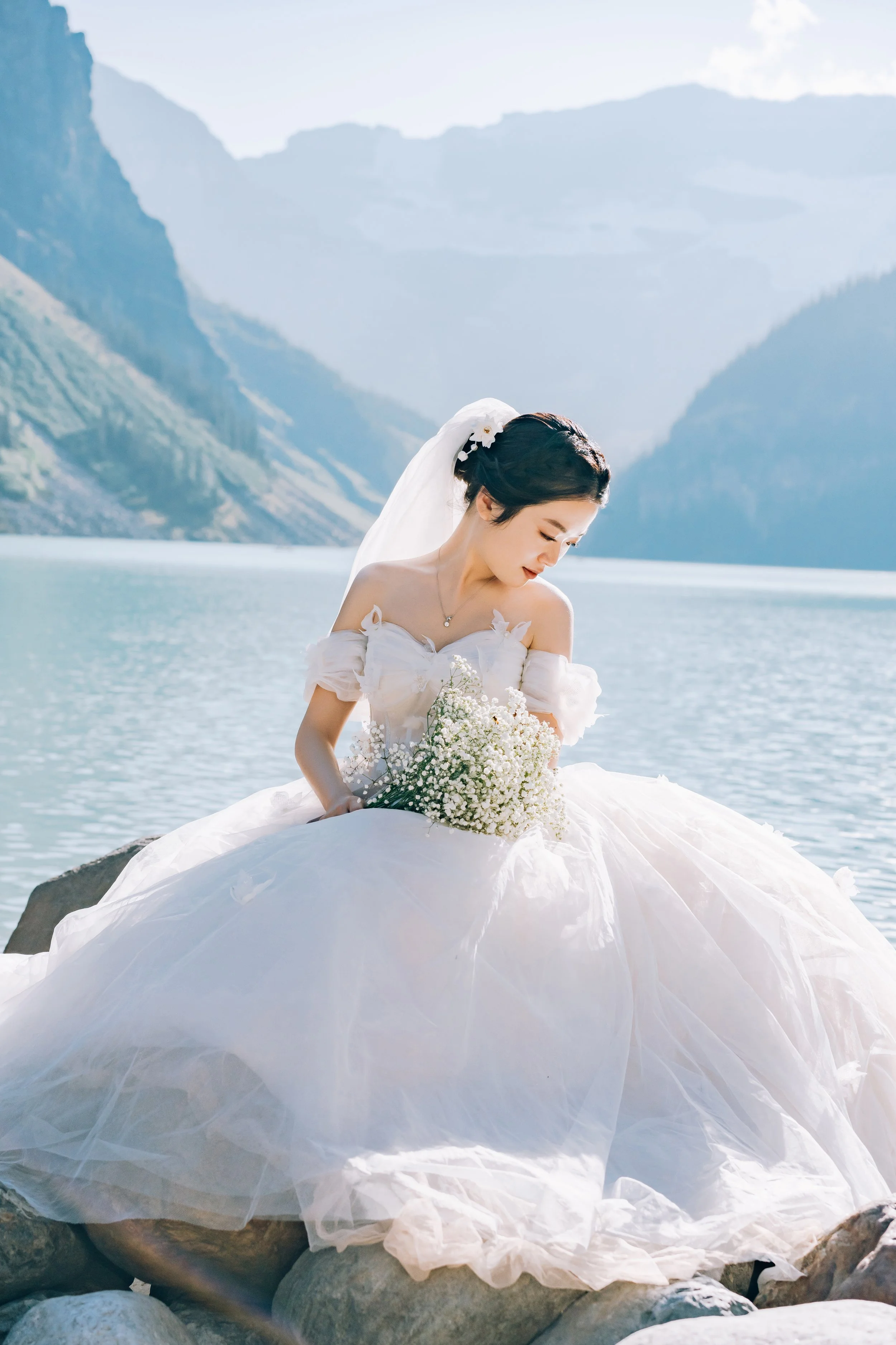 A bride sitting on rocks by a lake or river, wearing a white wedding dress, holding a bouquet of white flowers, with mountains and water in the background.