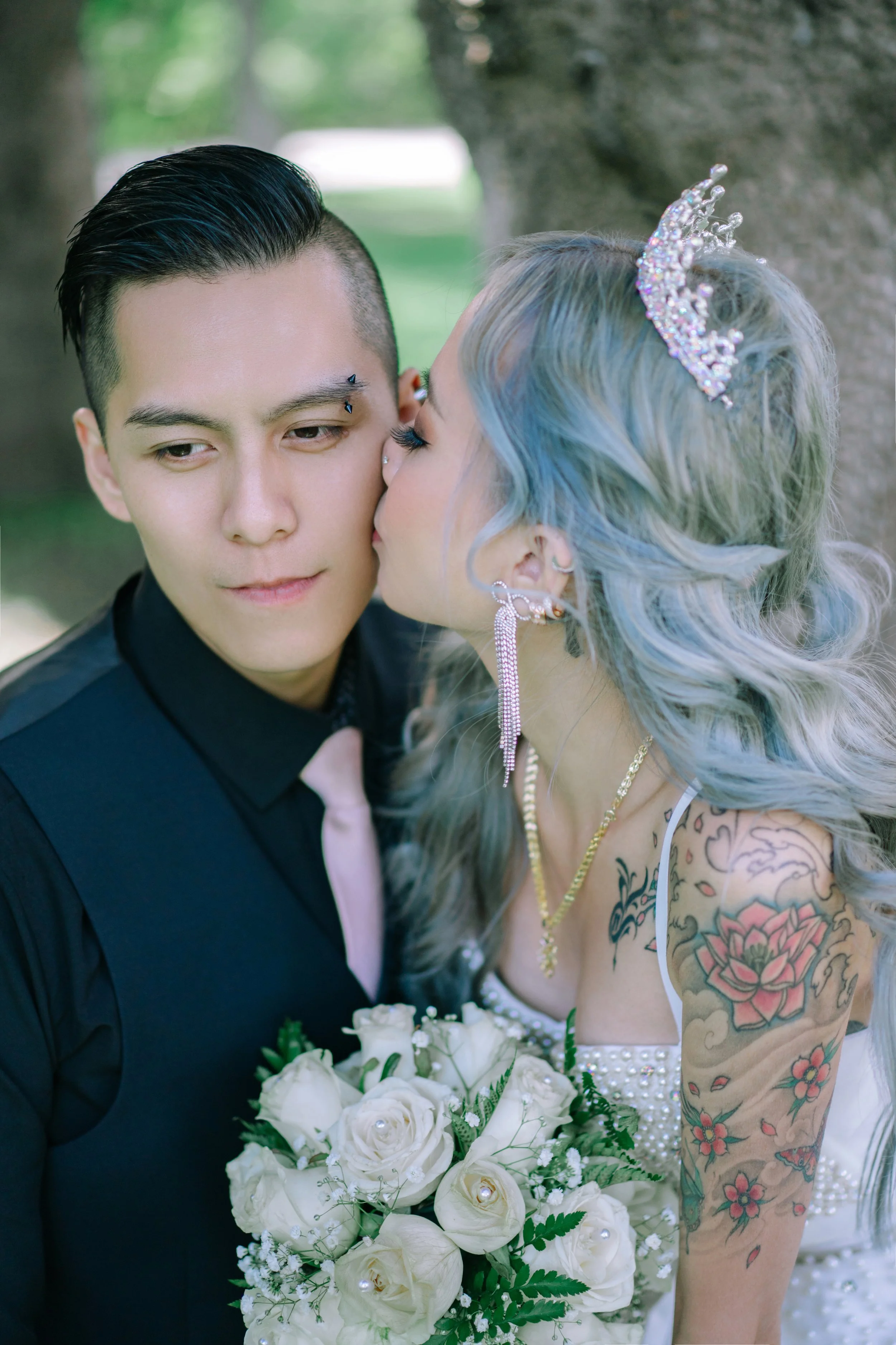 A woman with blue and silver hair wearing a tiara, earrings, necklaces, and a white dress, kisses a man with short black hair and an eyebrow piercing, wearing a black shirt and a pink tie, holding a bouquet of white roses, outdoors near trees.