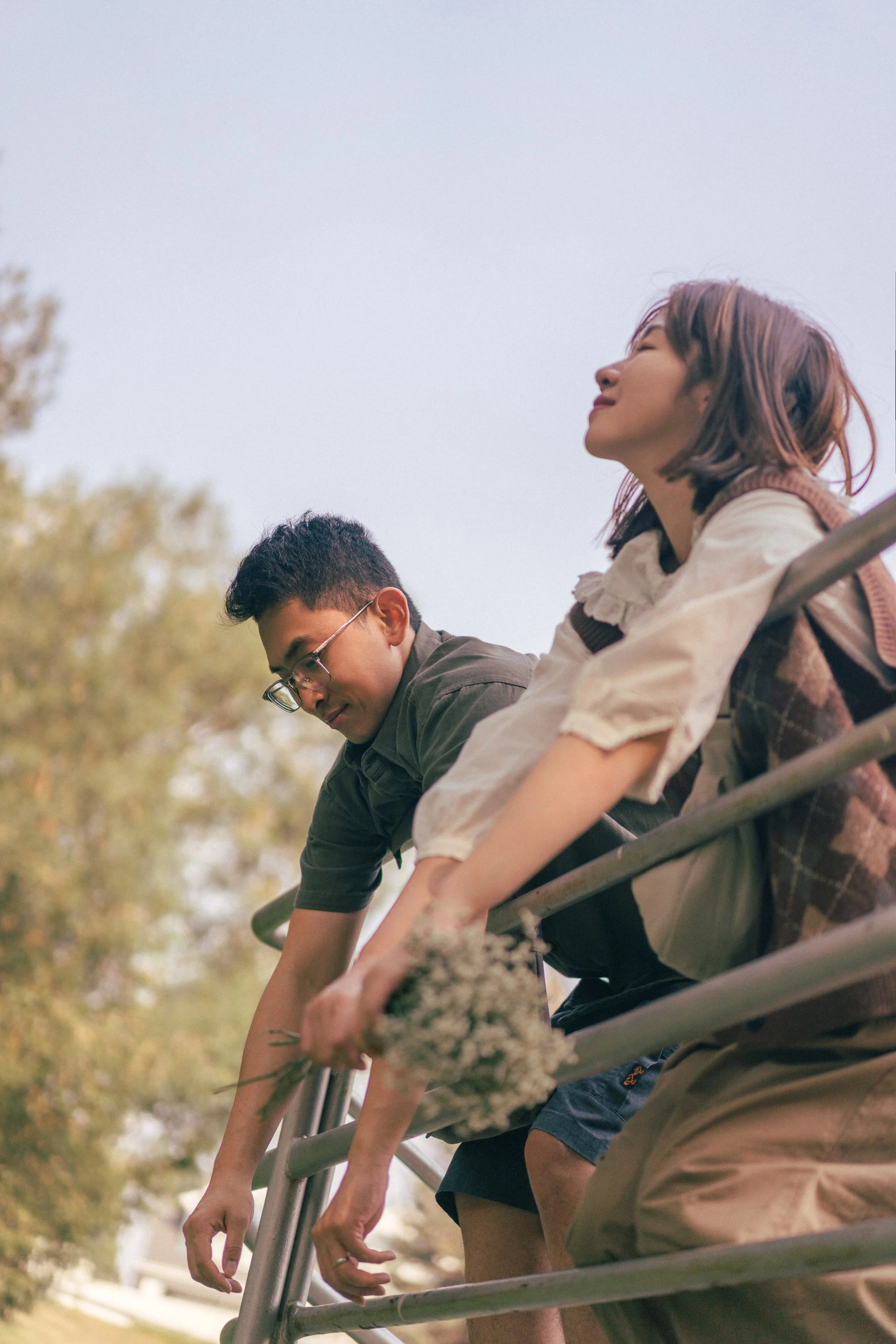 Two teenagers sitting on a metal railing outdoors, one holding a bunch of flowers, with trees in the background.