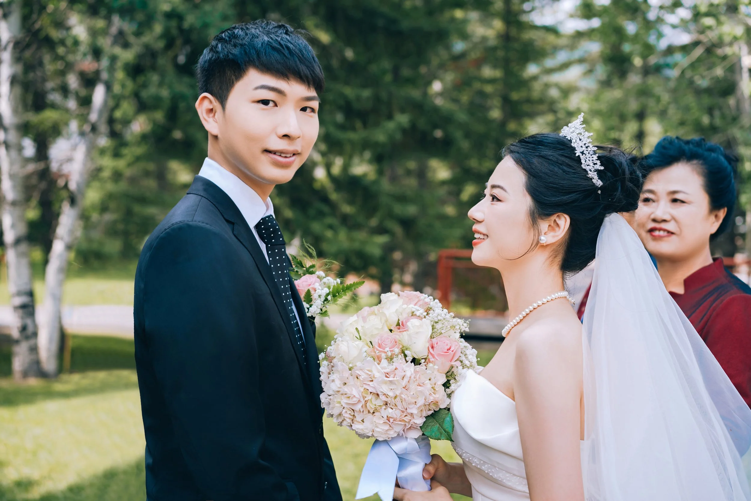 Bride in white wedding gown holding bouquet of pink and white roses, smiling at groom in black suit with tie, outdoors with greenery, woman in red dress in background.