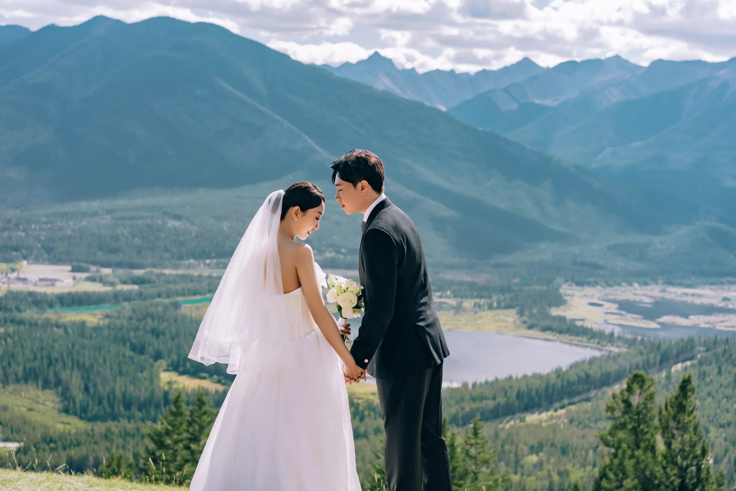 A bride and groom holding hands and looking at each other, standing outdoors with mountains, trees, and a lake in the background.