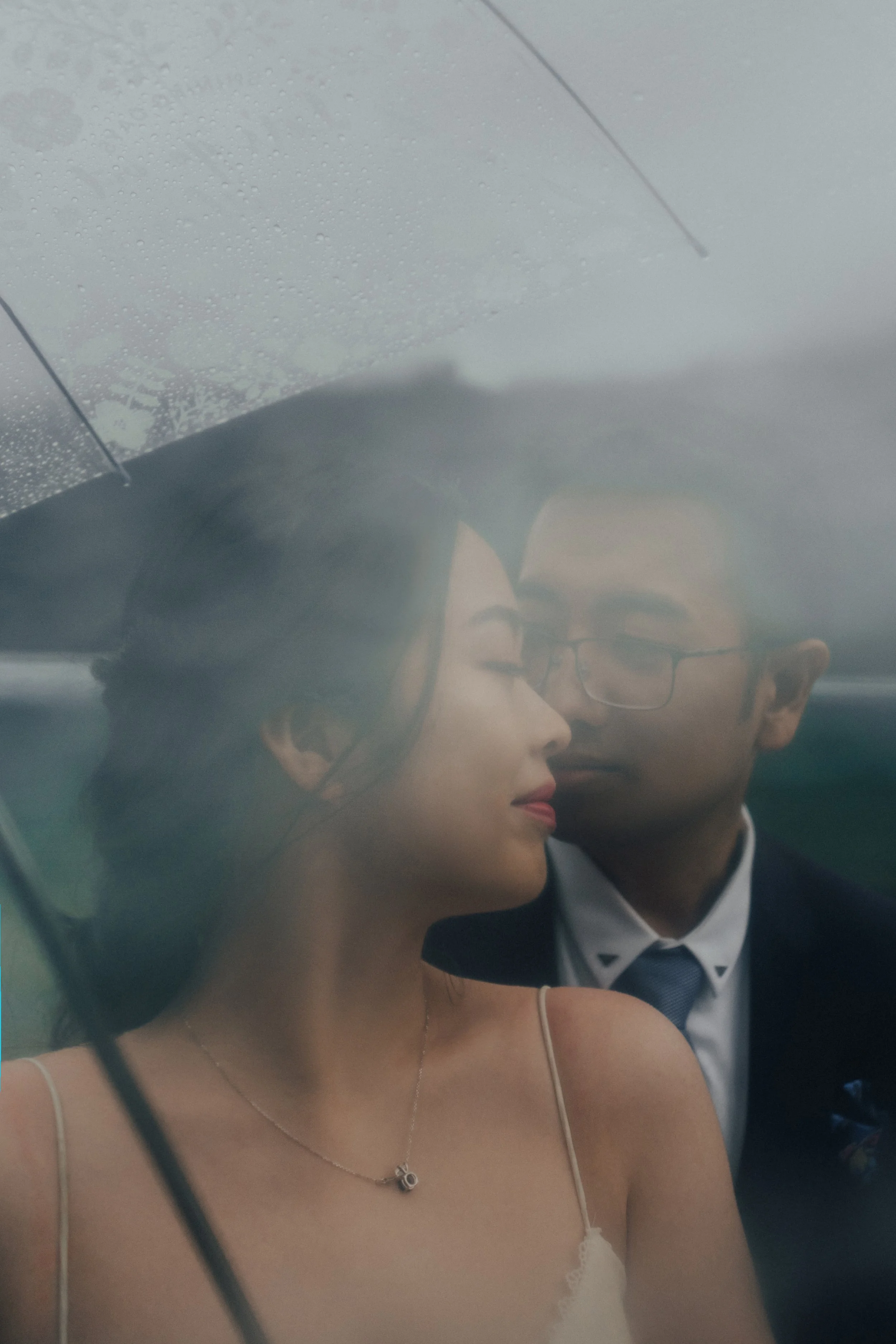 A couple sharing an intimate moment under a transparent umbrella with rain droplets, with a woman in a light-colored dress and a man in glasses and a suit, both with their eyes closed.