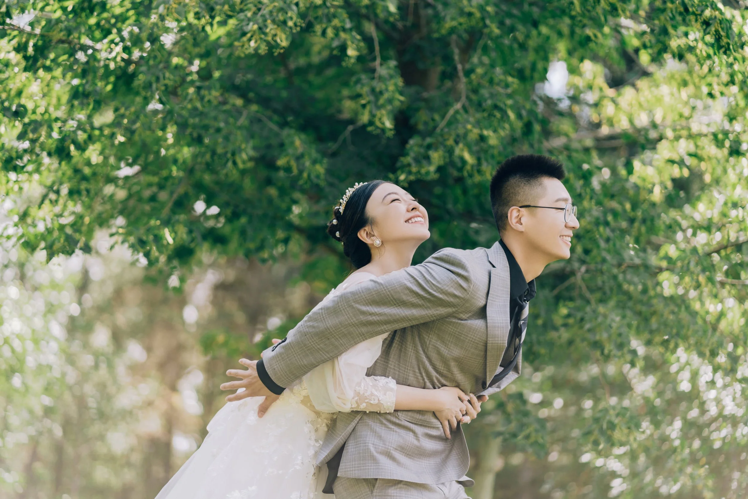 A joyful couple dressed in wedding attire stands outdoors under green trees, with the woman playfully leaning on the man's back.