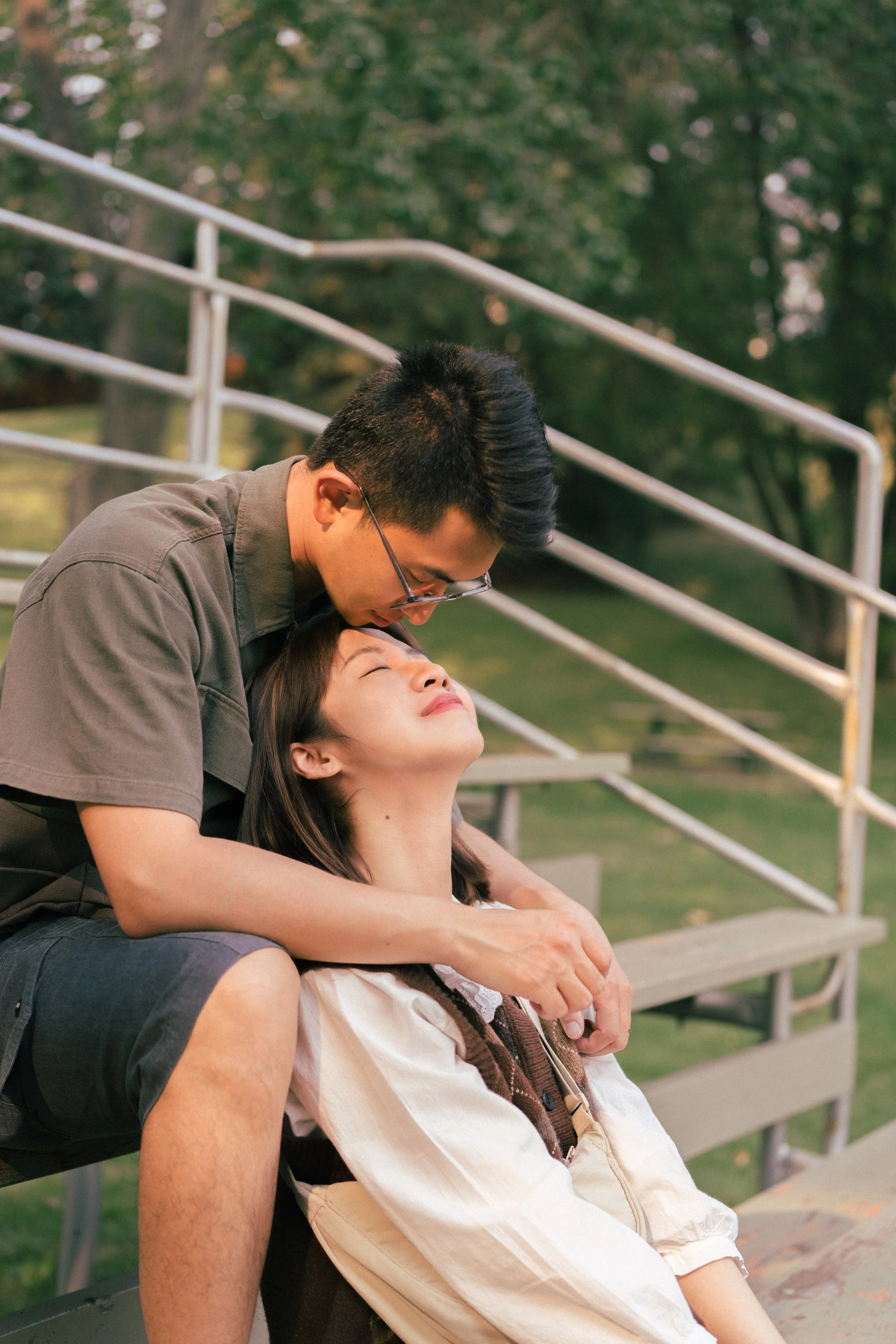 A young man leaning over a young woman sitting on a park bench, gently touching her face and forehead. They are outdoors with green trees and metal railings in the background.