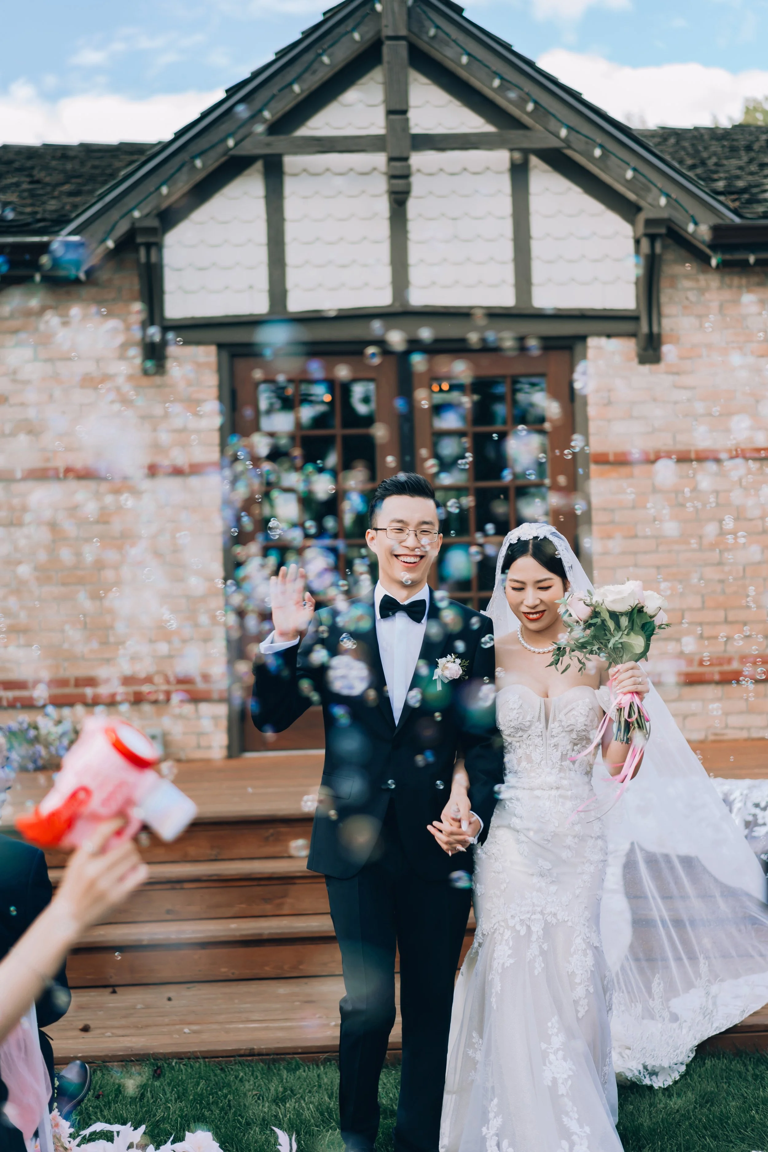A newlywed couple in wedding attire holding hands and smiling outside a brick house with a wooden staircase and a window, surrounded by bubbles.