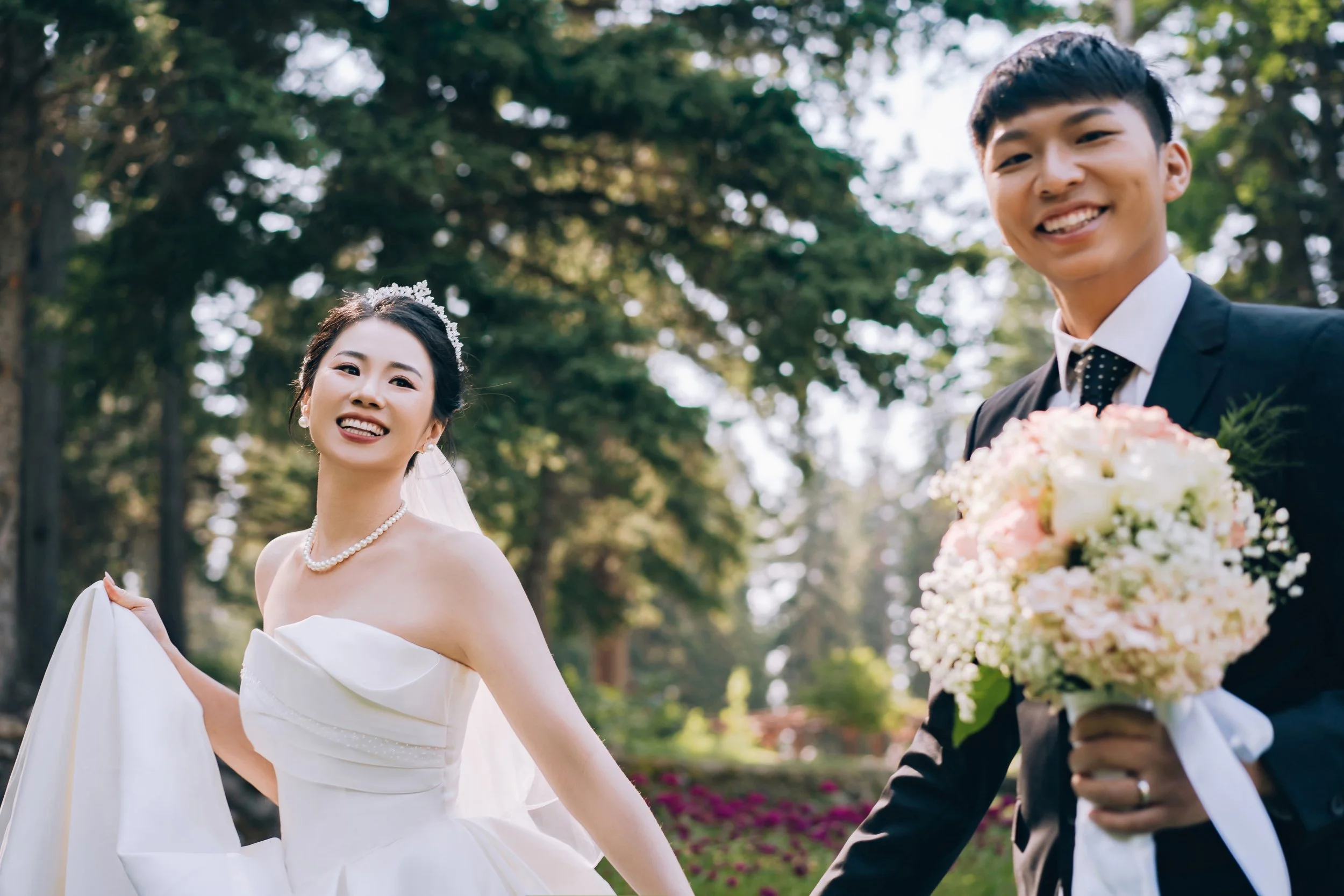 A bride and groom smiling outdoors on their wedding day, with trees in the background. The bride wears a white strapless wedding gown, pearls, and a tiara, while the groom, in a suit and tie, holds a large bouquet of flowers.