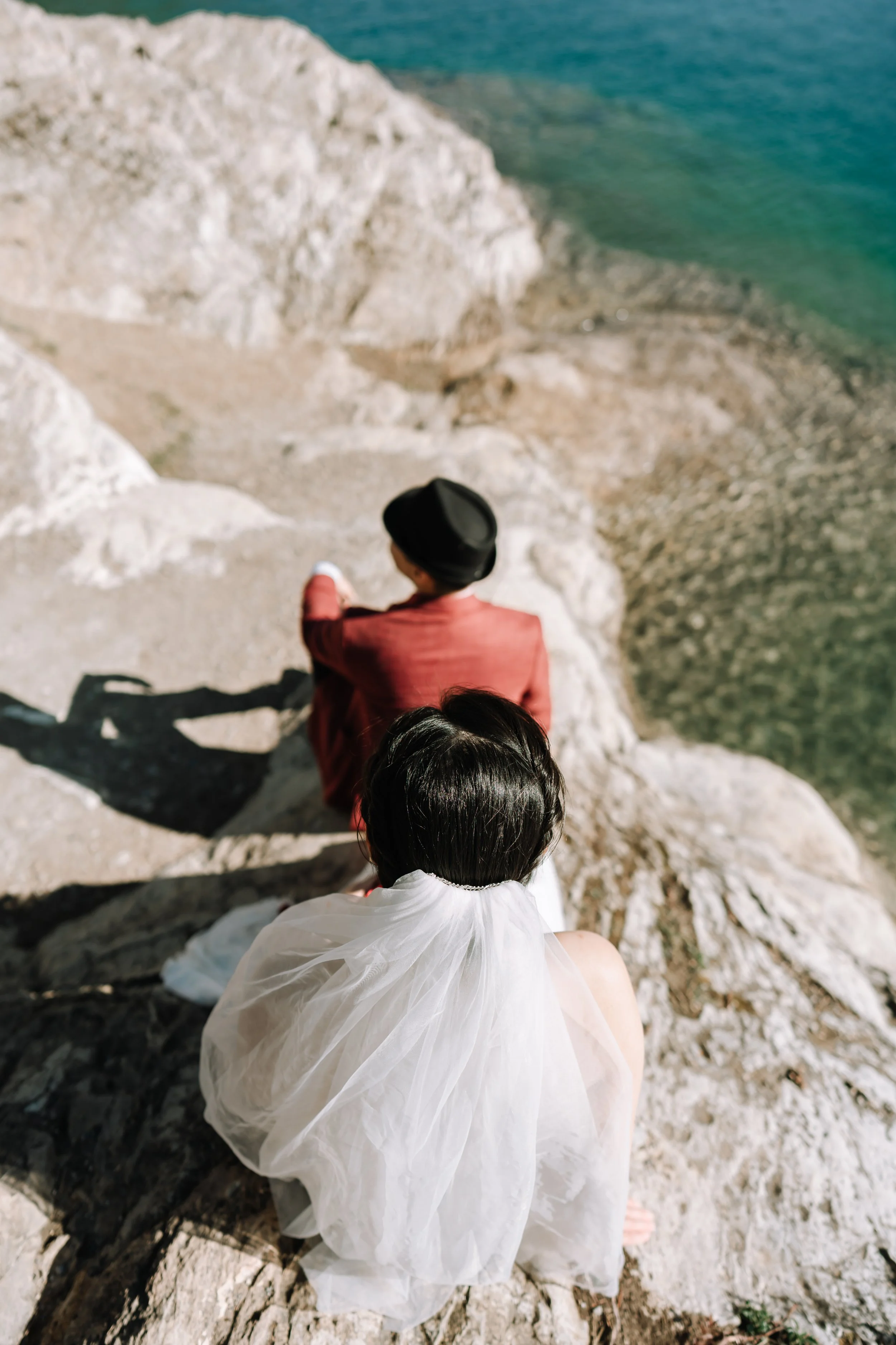 A woman with short black hair wearing a white dress and veil sitting on a rocky cliff, with a man in a red jacket and black hat sitting nearby, overlooking a coastal scene with rocks and clear blue water.