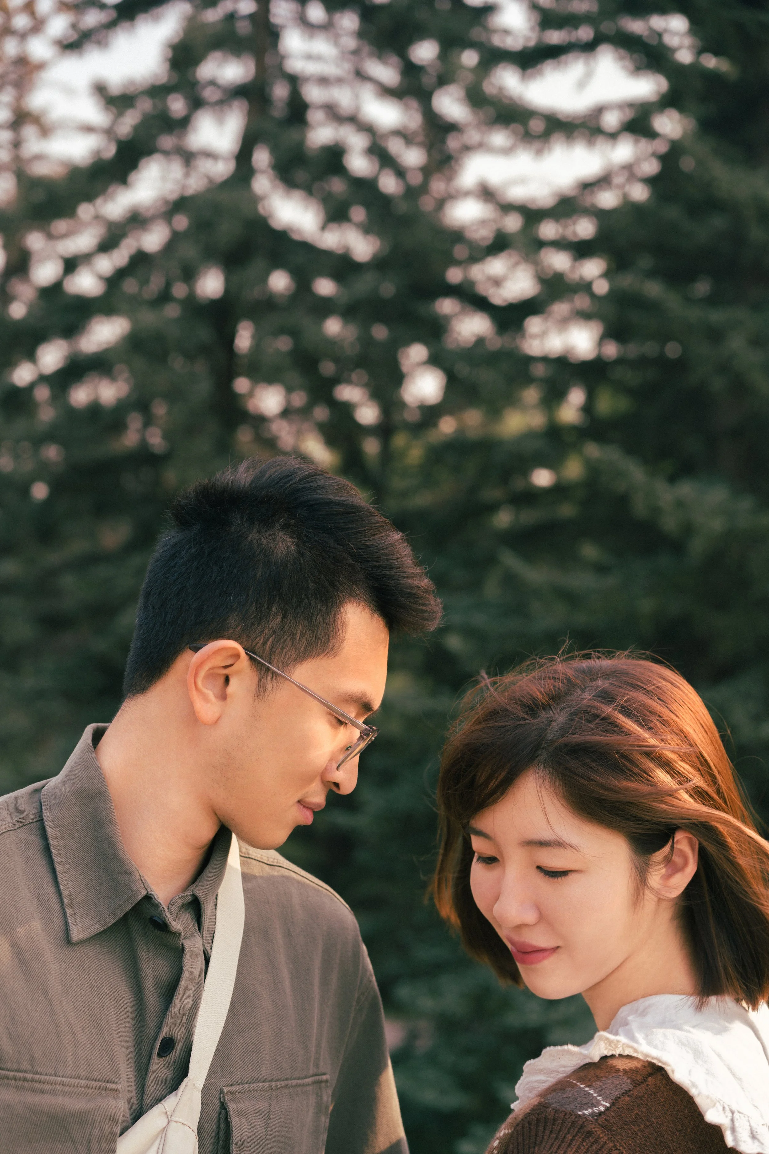 A young man and woman standing close together outdoors with trees in the background during sunset.