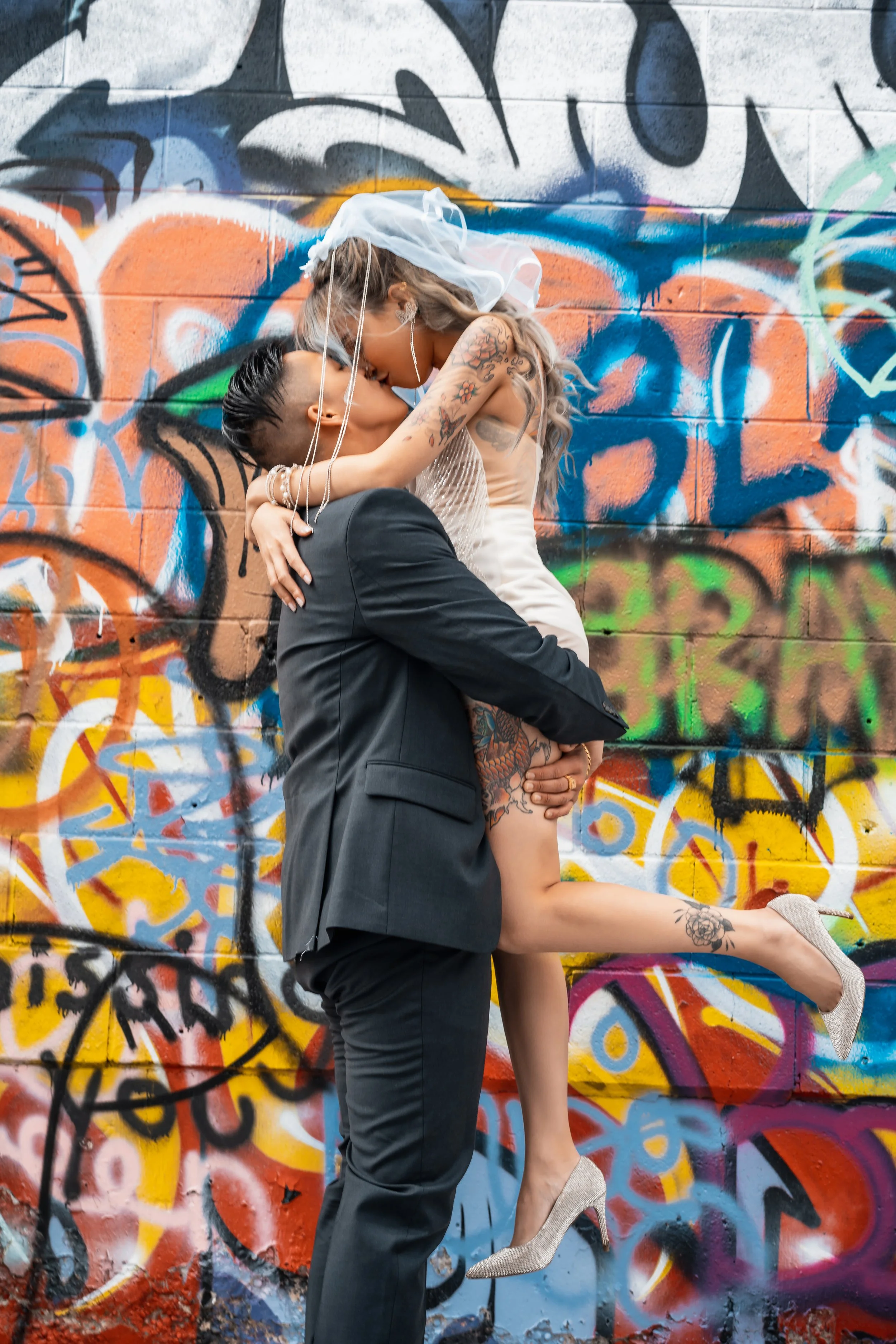Bride and groom kissing in front of colorful graffiti wall, man in suit holding woman in wedding dress and veil.