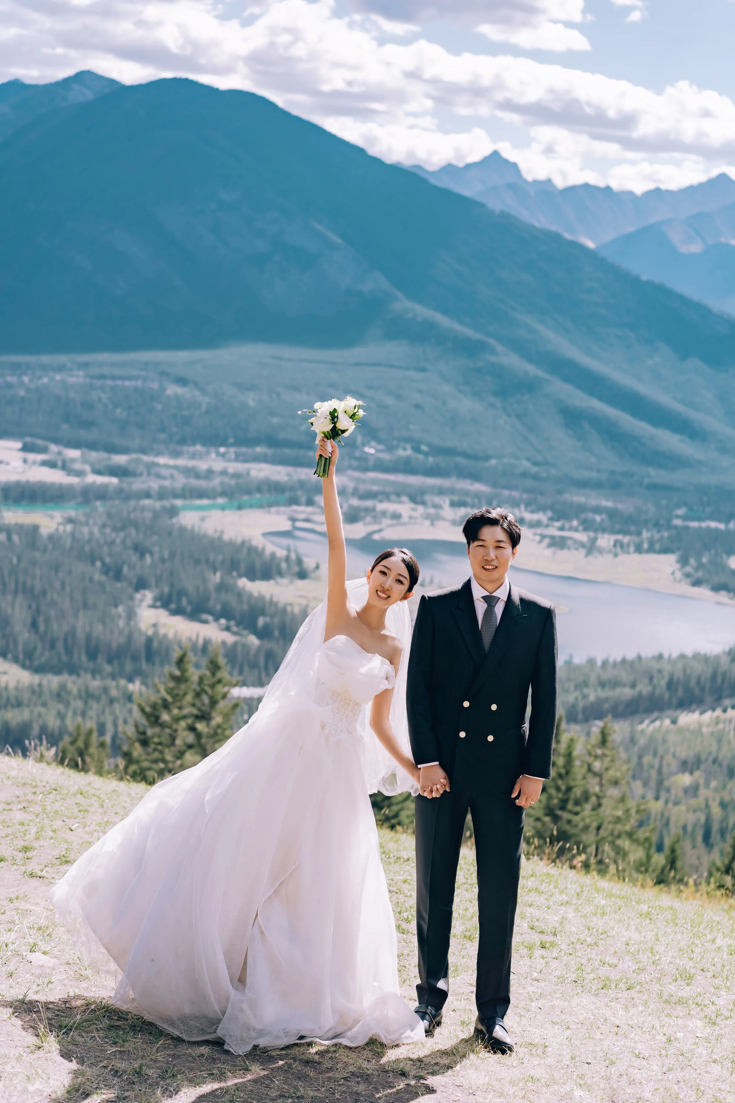 A bride and groom holding hands outdoors on a mountain overlook, with a scenic mountain range, lake, and forest in the background. The bride is wearing a white wedding gown and holding a bouquet of white flowers, raising her arm celebrating. The groo