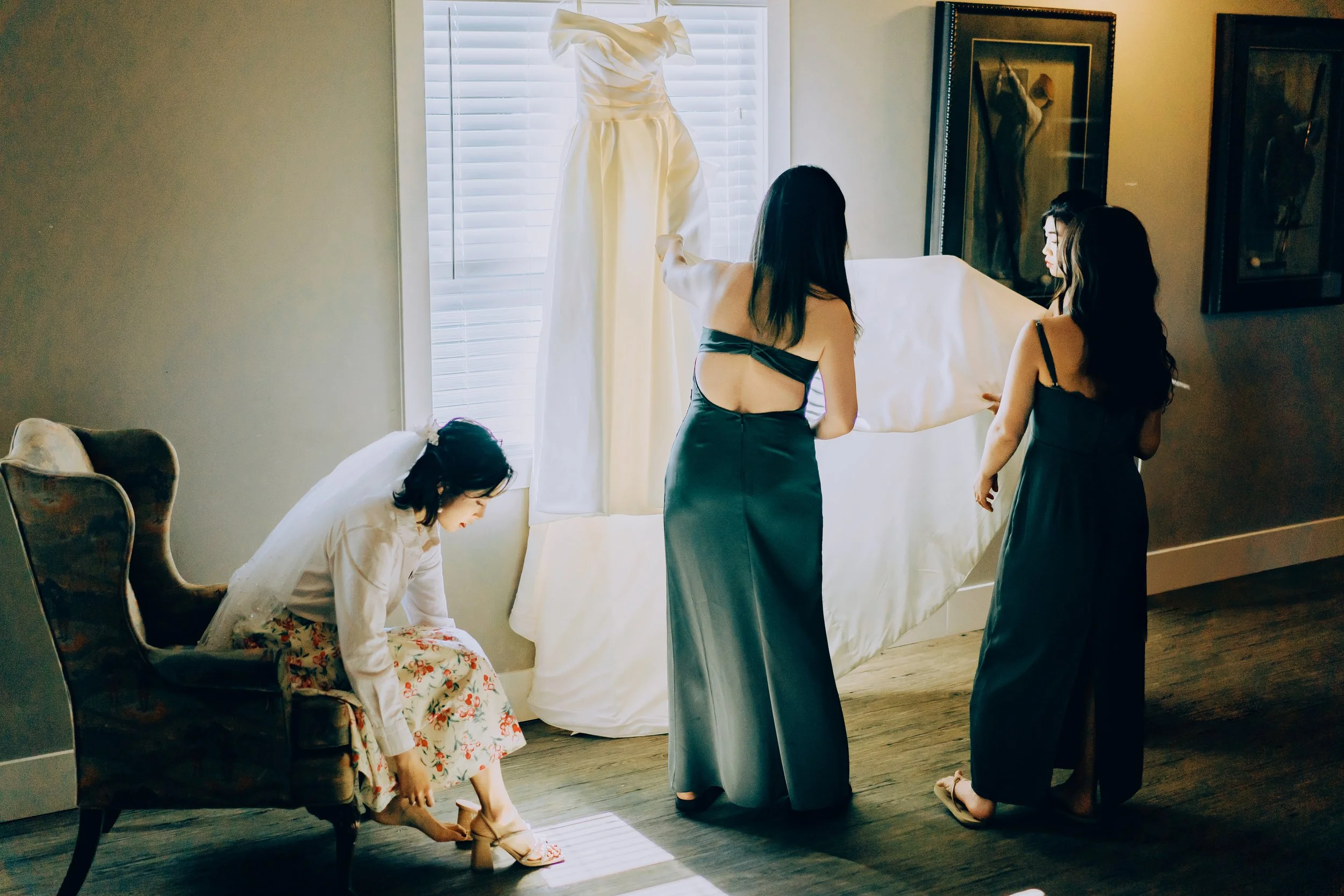 Bridal party preparing in sunlight, bride adjusting shoes, wedding dress displayed on window, bridesmaids in teal dresses.