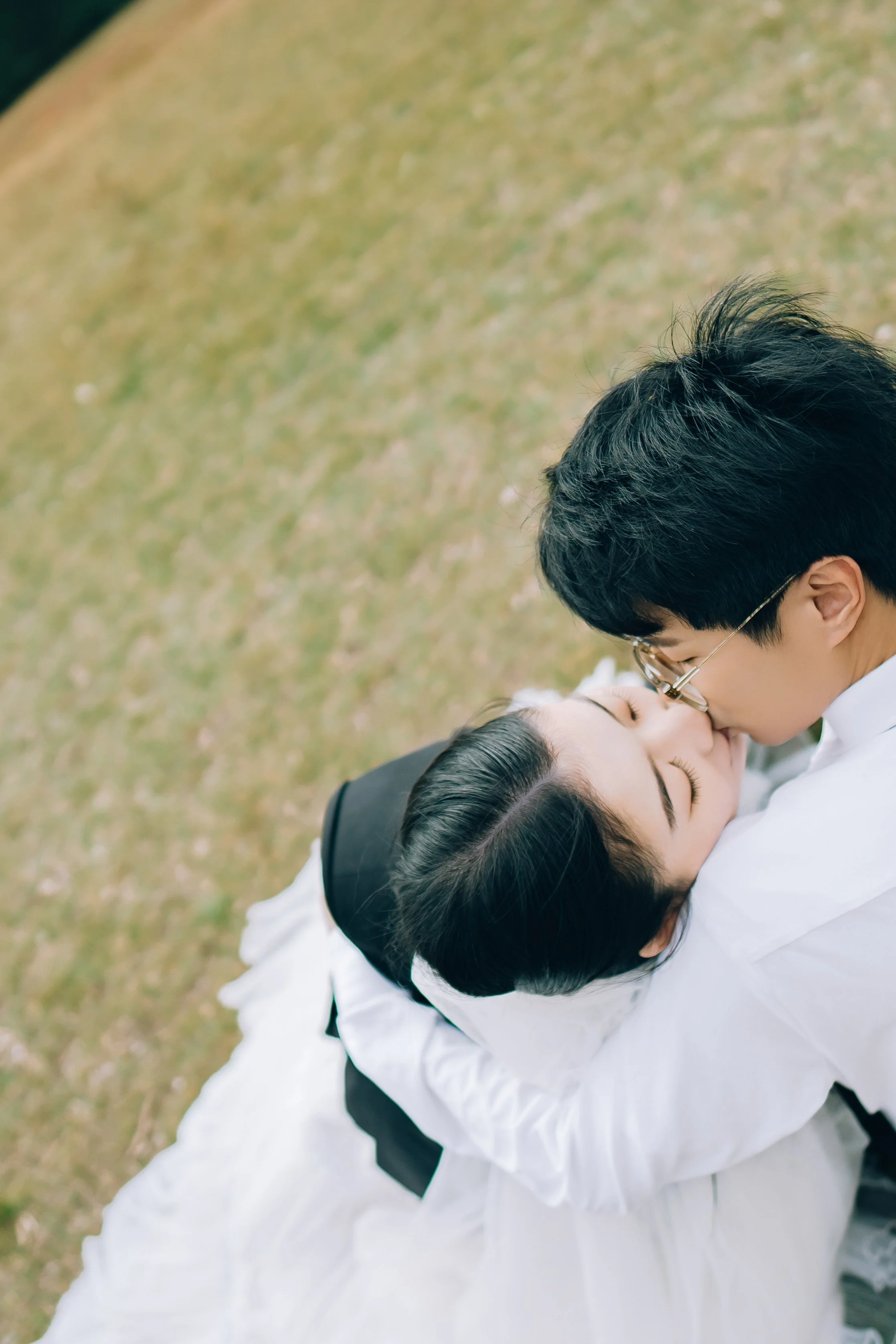 A young couple in wedding attire sharing a kiss outdoors on a grassy area.