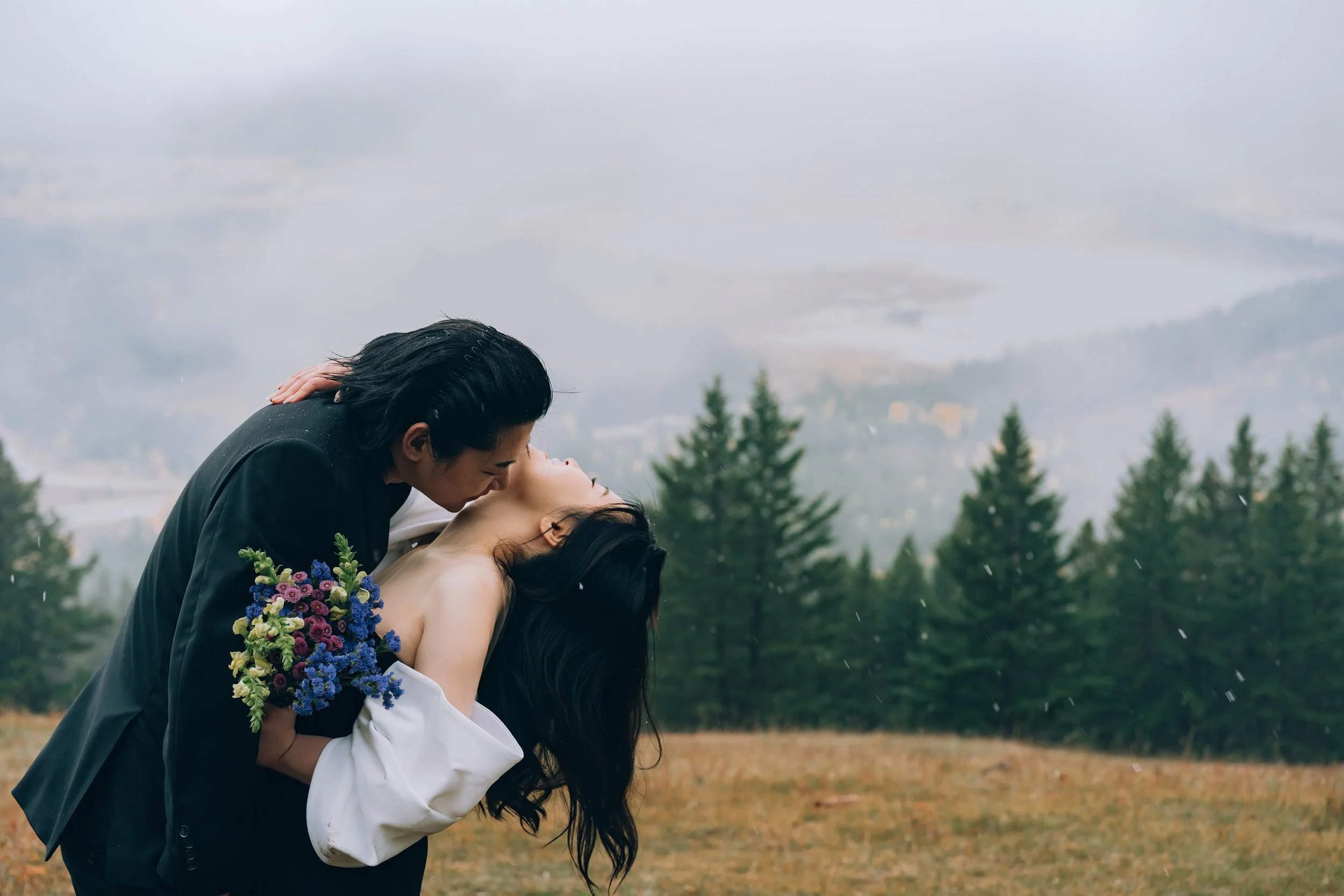 A couple kissing outdoors in a rainy, mountainous landscape, with a woman holding a colorful bouquet of flowers.