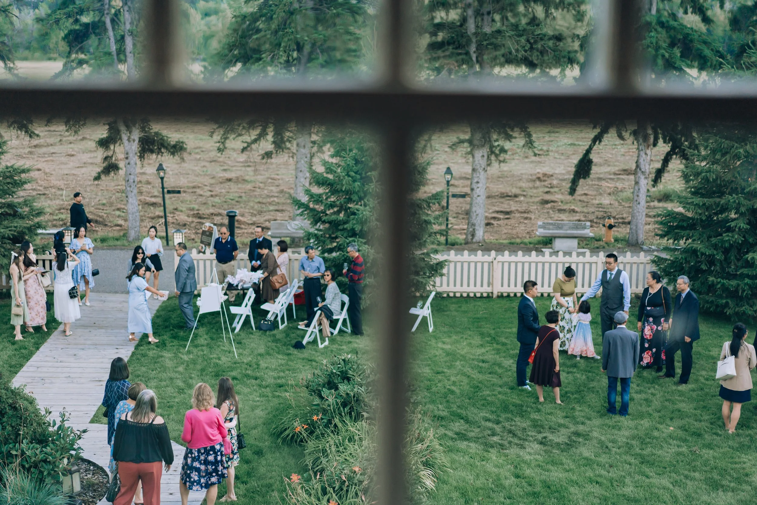 People gathered in an outdoor garden, possibly for a wedding or celebratory event, seen through a window with a cross-shaped grid.