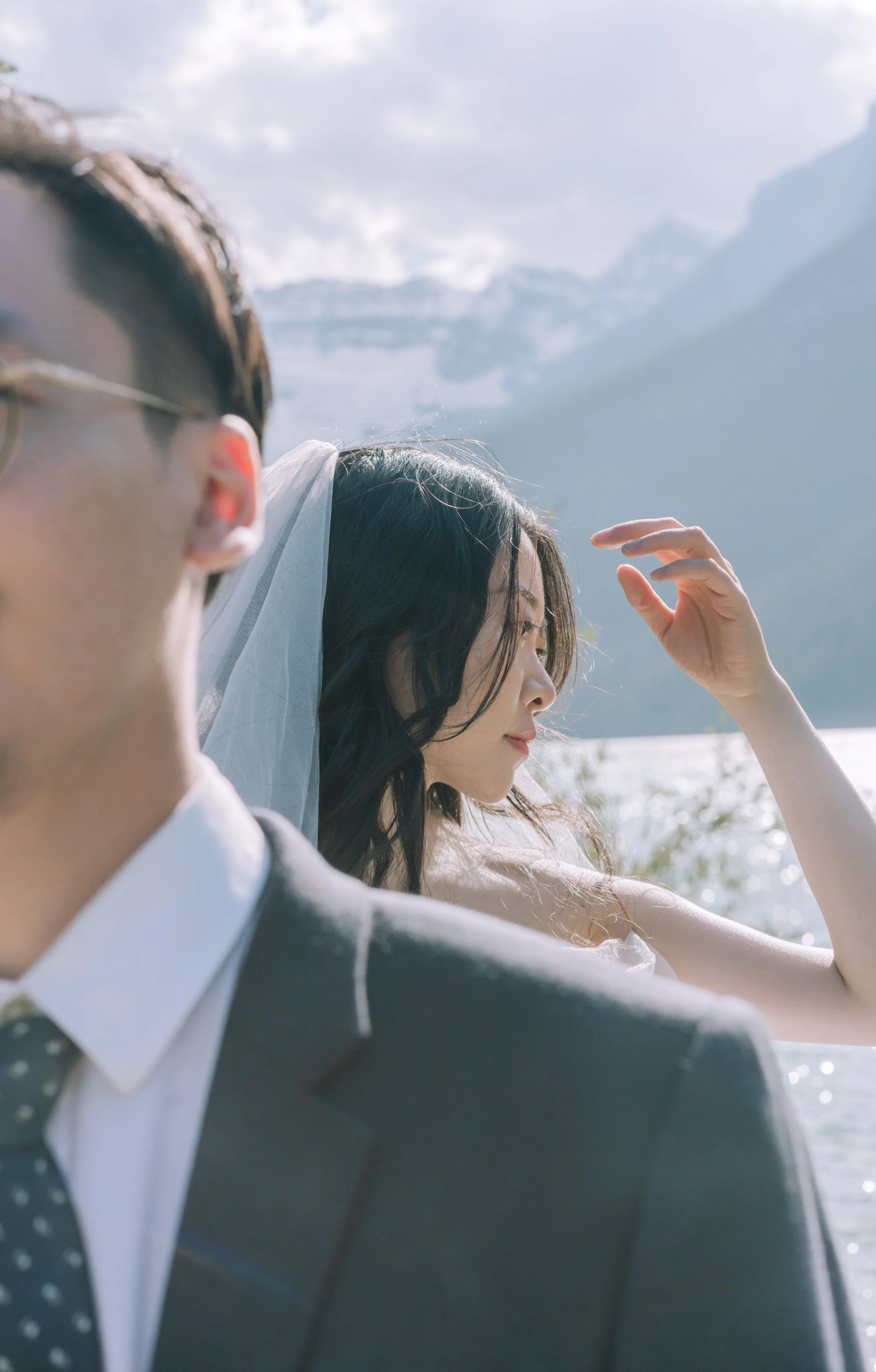A bride with dark wavy hair and a veil, and a groom in a suit with glasses, standing outdoors near a body of water with mountains in the background.