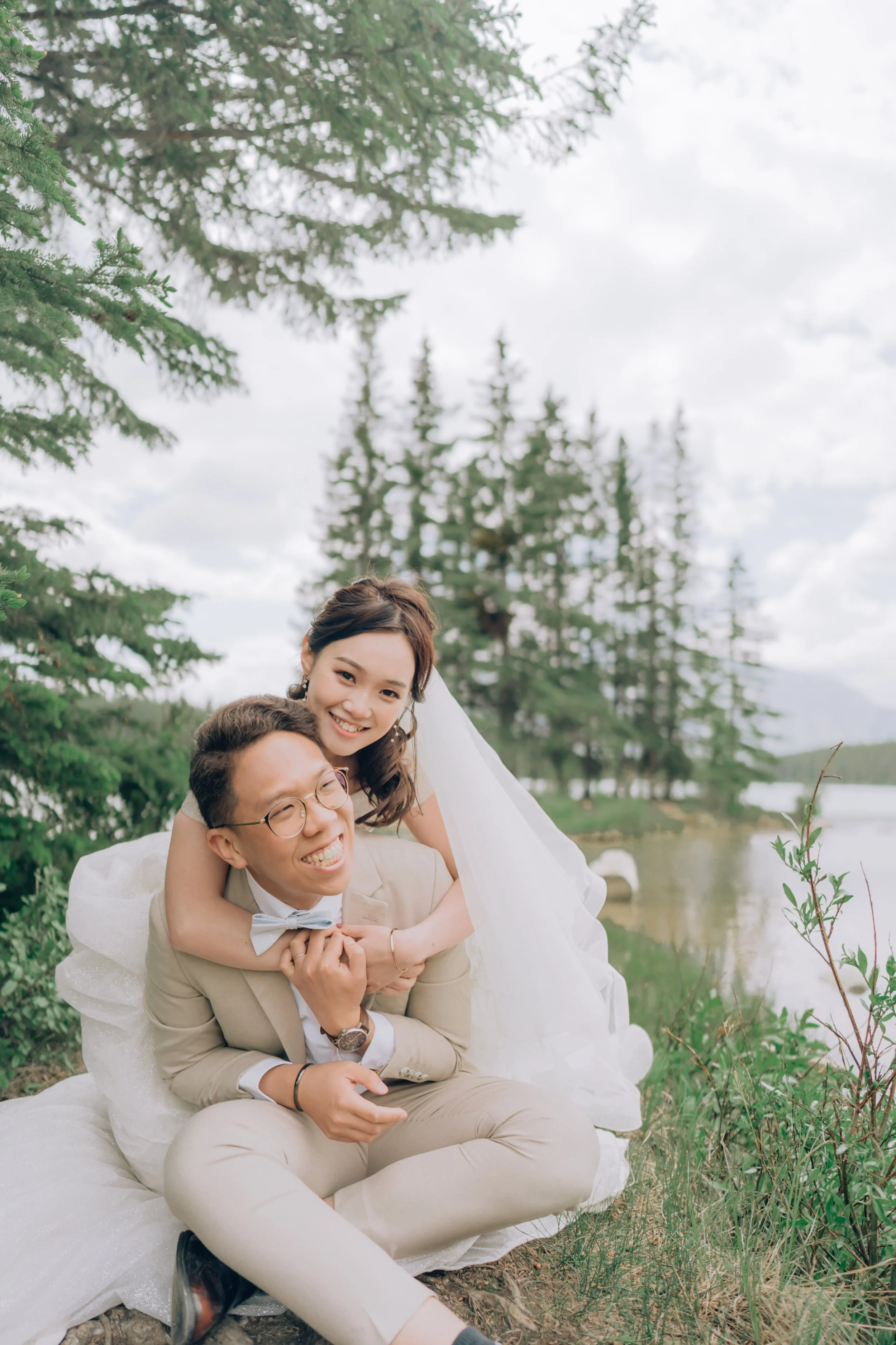 A happy bride and groom outdoors near a lake, surrounded by pine trees, with the bride leaning on the groom's shoulders and both smiling.