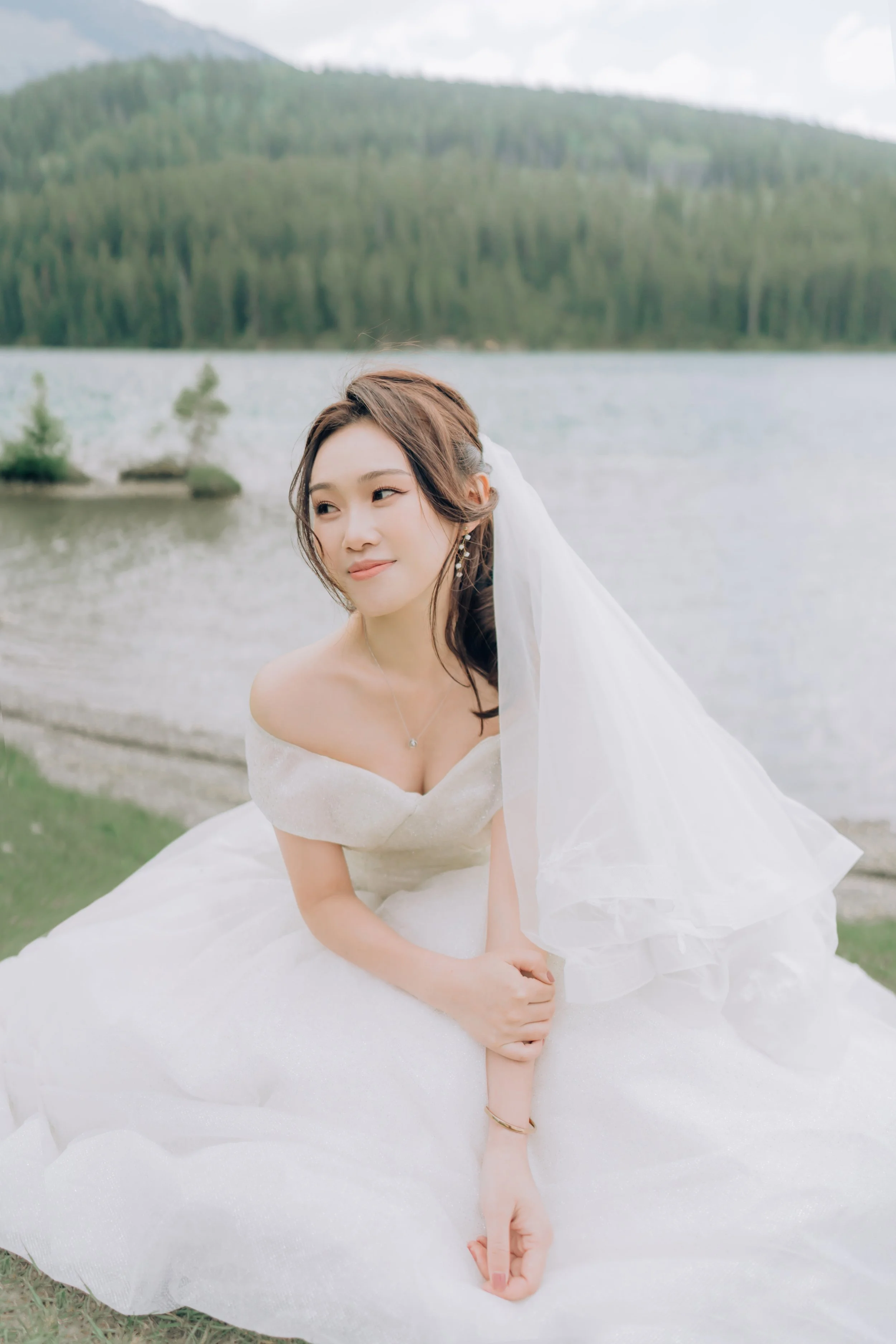 A bride in a white wedding dress sitting on grass near a lake with mountains and forest in the background.