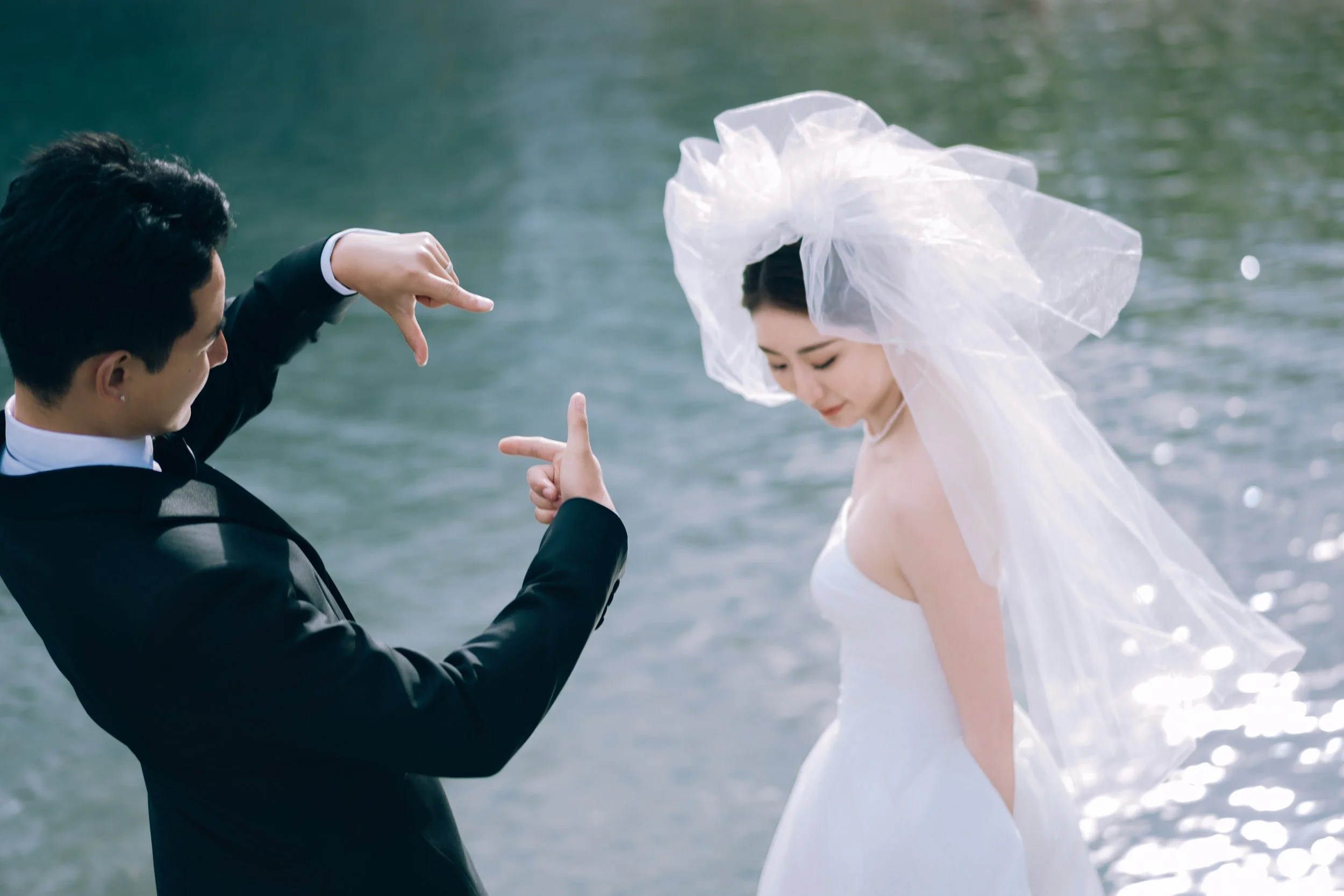 A groom in a black suit taking a photo of a bride in a wedding dress near a body of water.