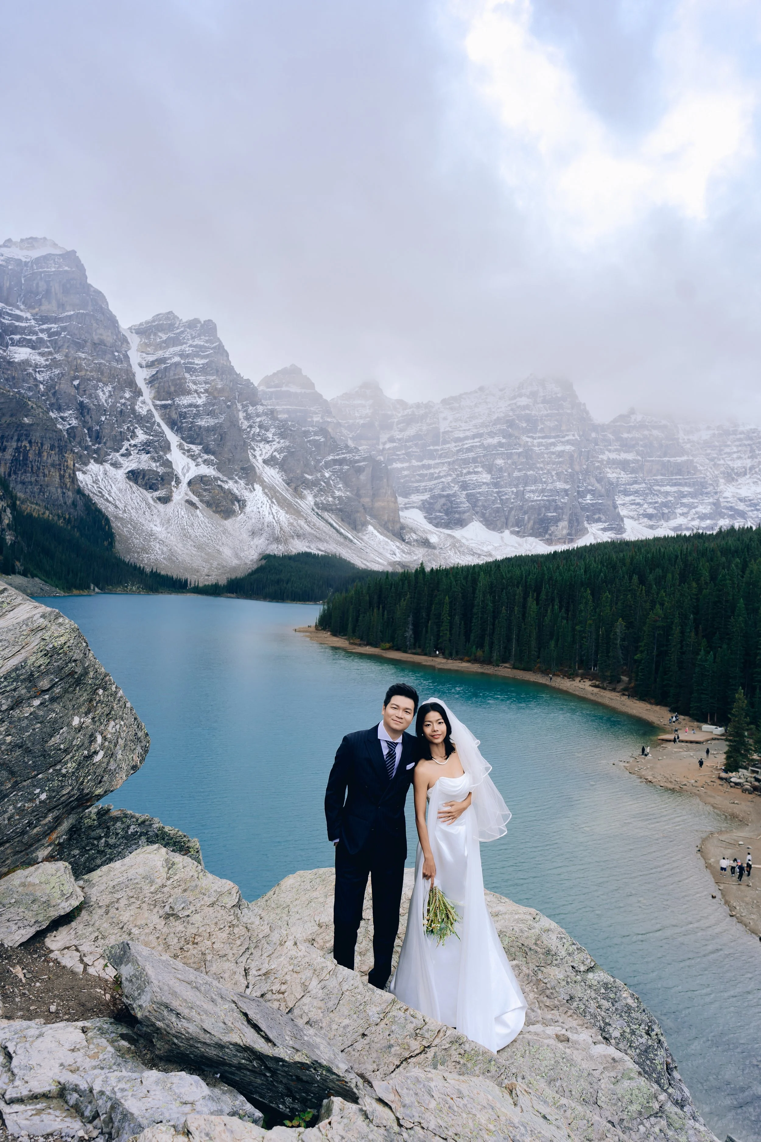 A bride and groom standing on rocks by a lake with mountains and forests in the background.