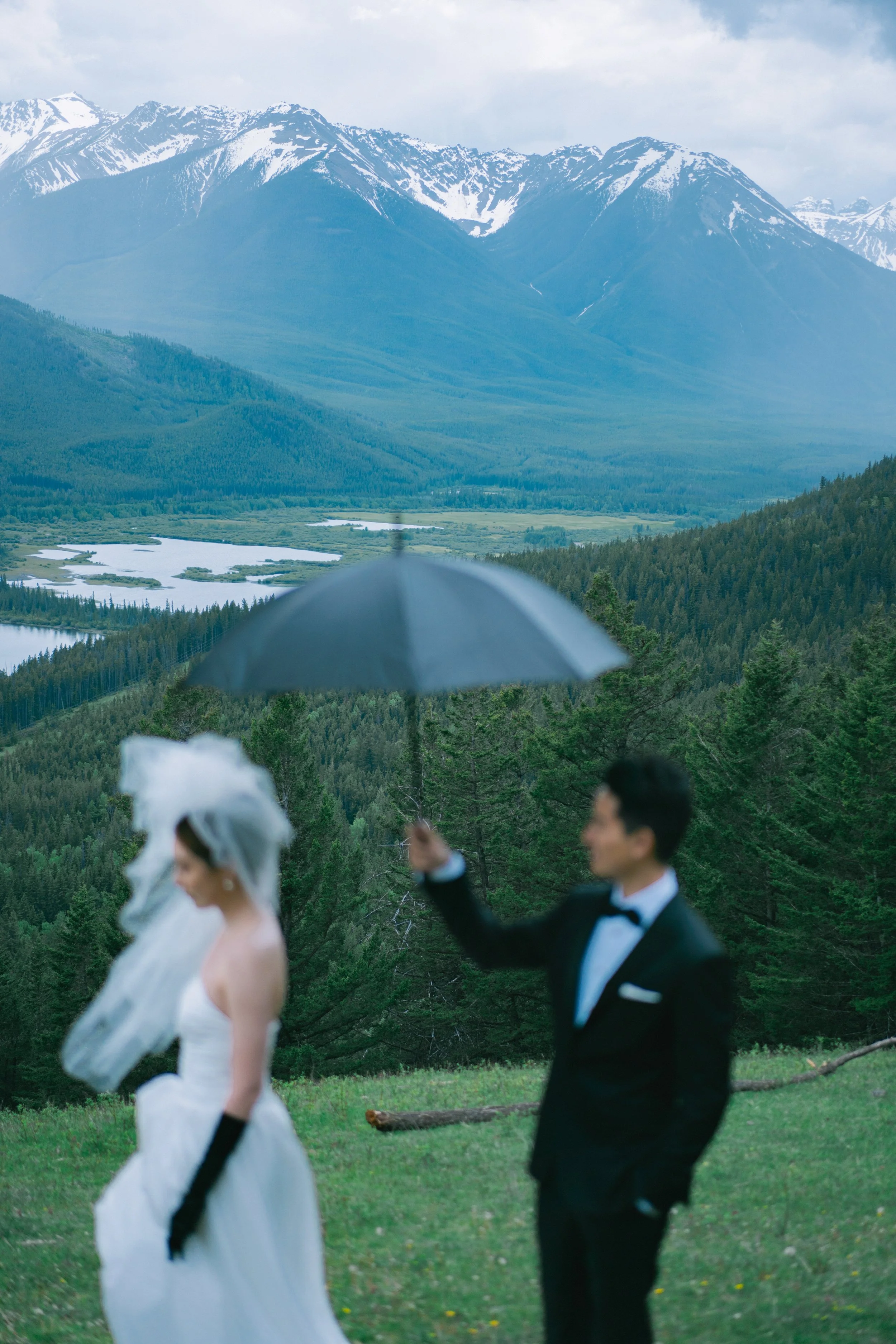 A bride and groom dressed in wedding attire standing outdoors in a green field with mountains and a lake in the background. The groom holds a black umbrella over the bride.