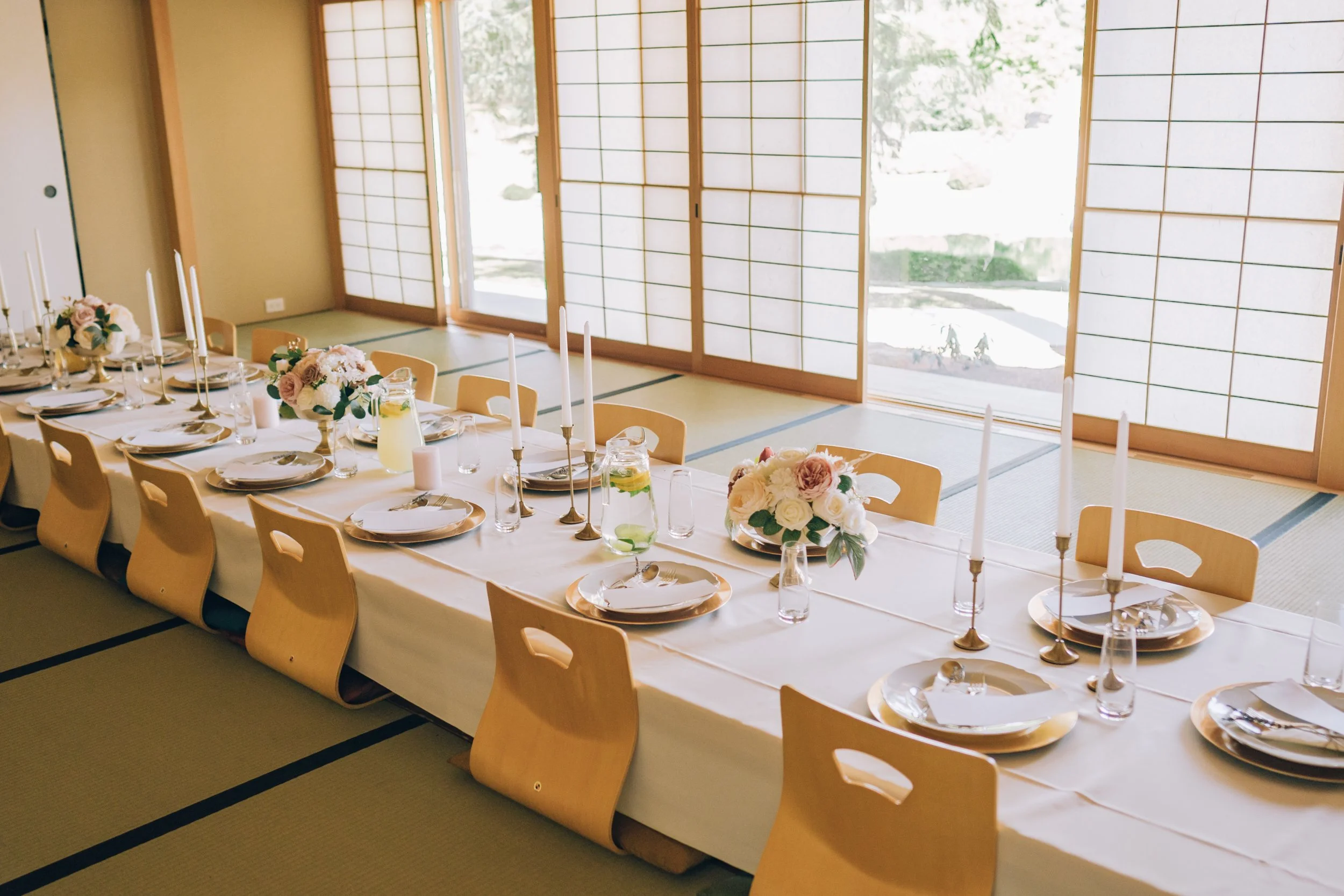 Elegant indoor dining table set with floral centerpieces, white plates, candles, and cutlery, in a room with shoji screens and tatami mats.