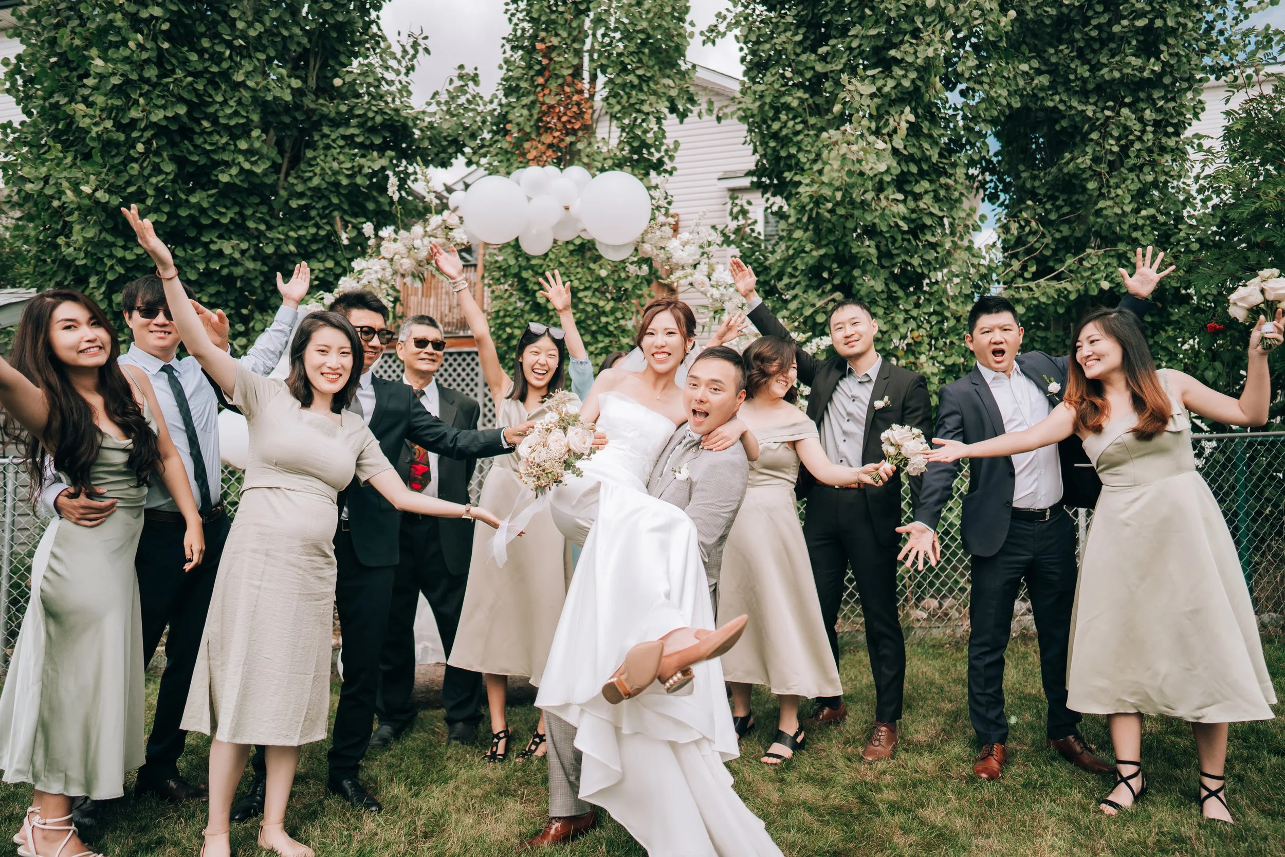 A wedding celebration outdoors with a bride and groom surrounded by friends and family, smiling and posing with arms raised, lush green trees in the background.