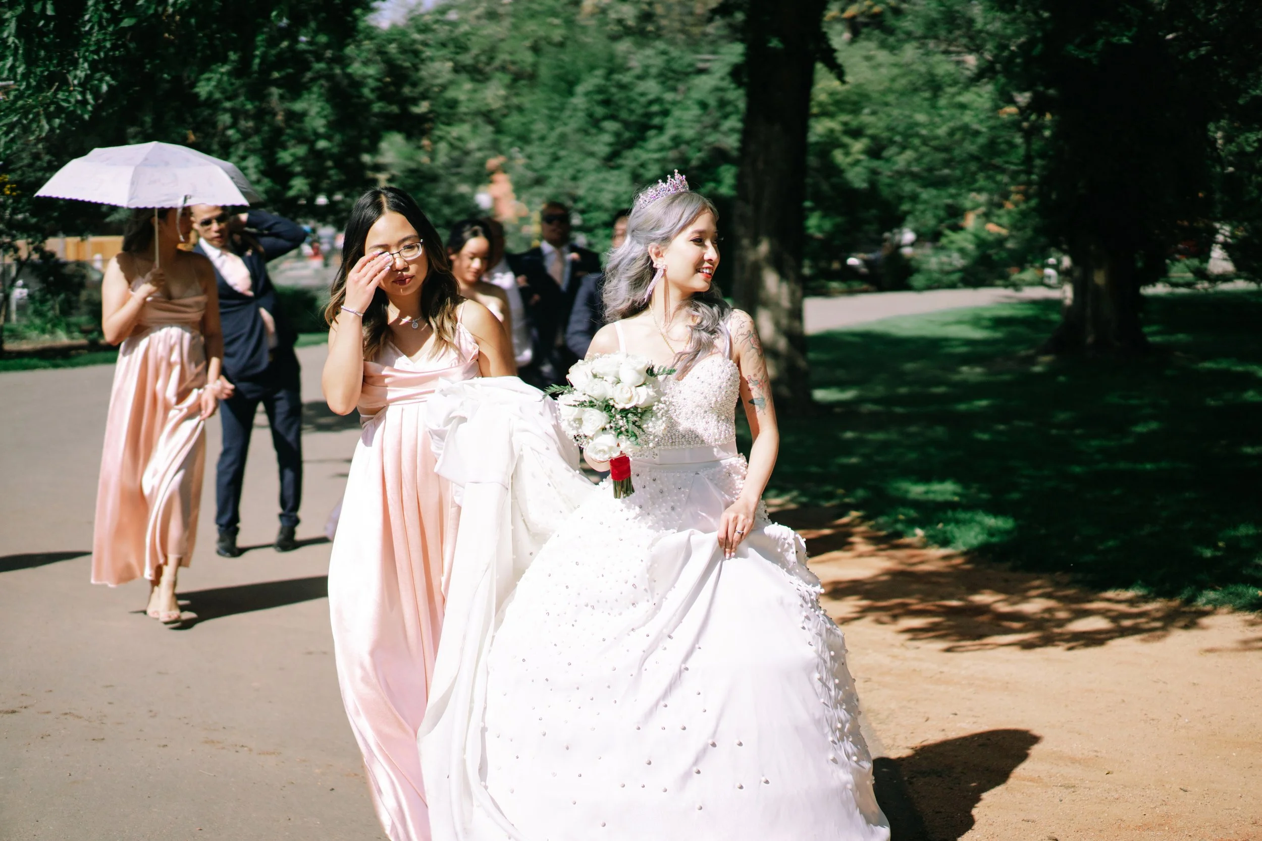 A bride in a white wedding gown with a bouquet of white roses walking outdoors, followed by bridesmaids and groomsmen, in a lush green park on a sunny day.