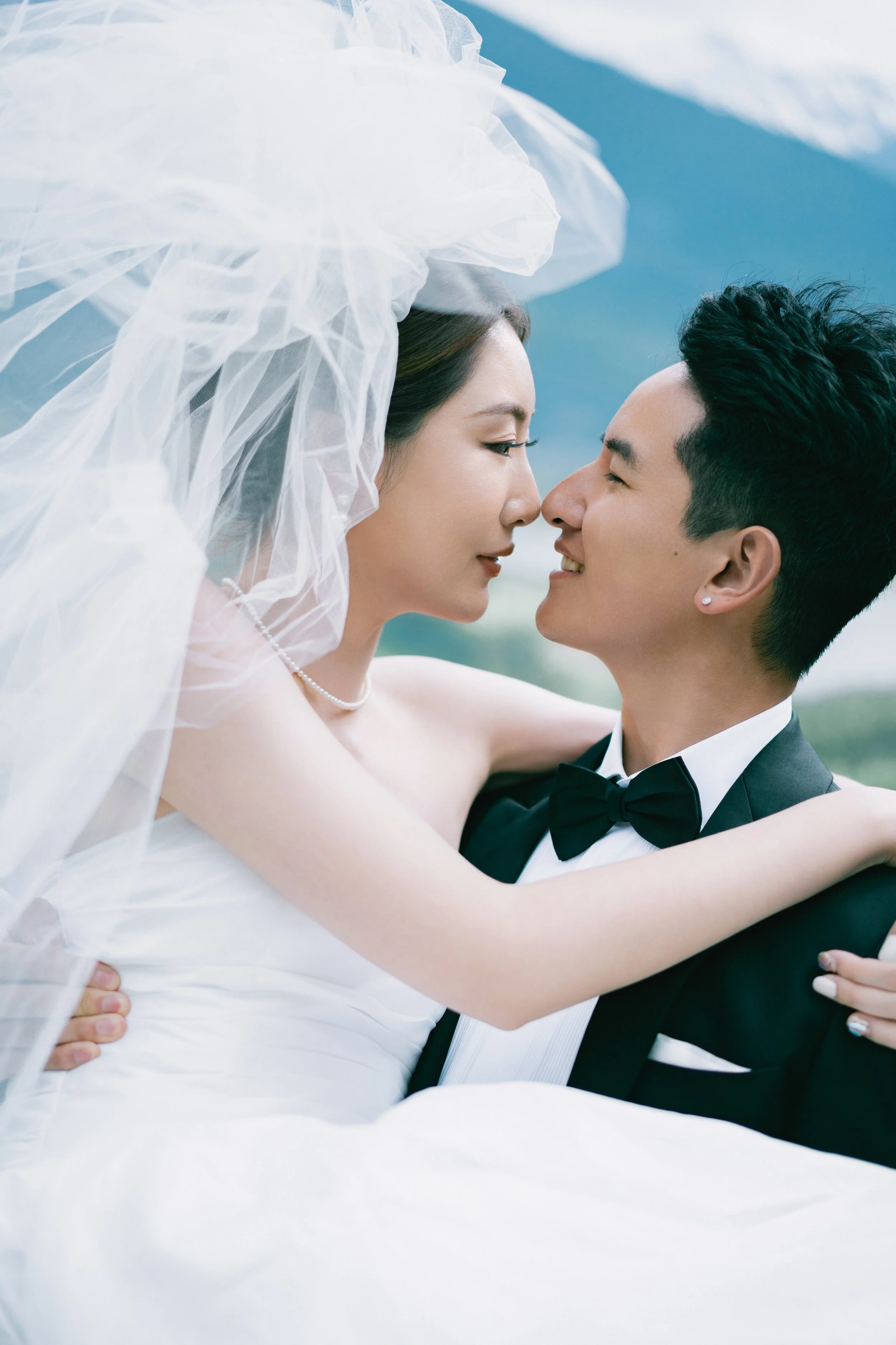 A bride and groom in wedding attire embracing outdoors, with their faces close together and smiling.