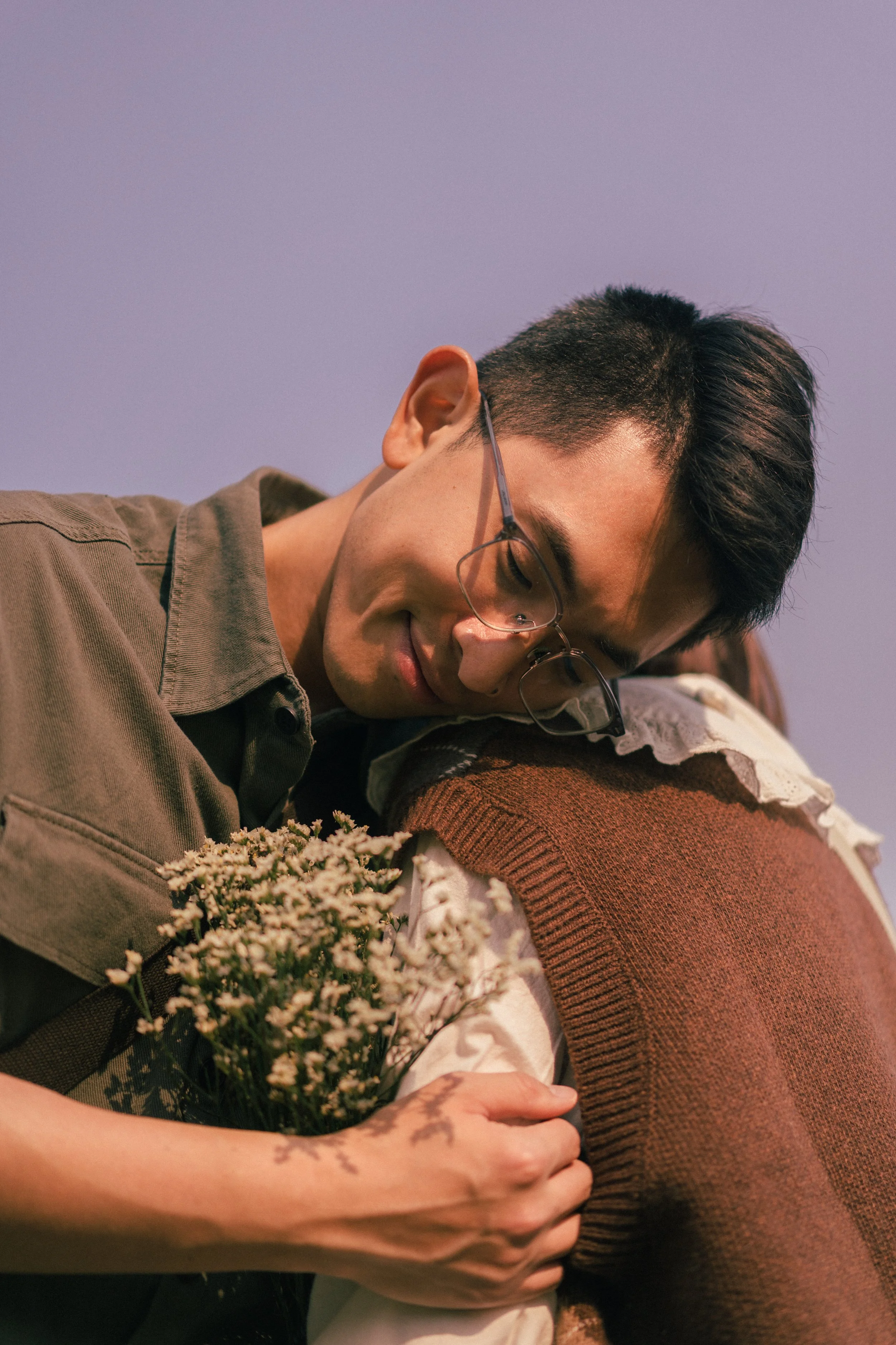 A person with short dark hair and glasses resting their head on another person's shoulder, holding a bouquet of small white flowers. The background features a clear sky.