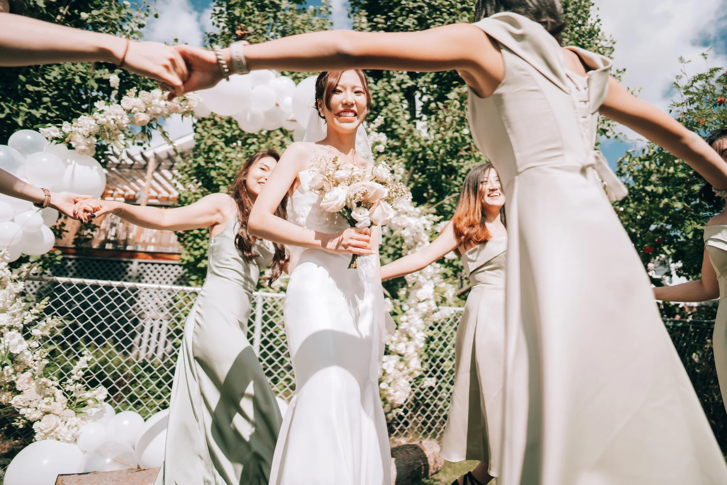 A bride holding a bouquet of flowers, standing in a circle with bridesmaids during an outdoor wedding ceremony, with white balloons and floral arch in the background.