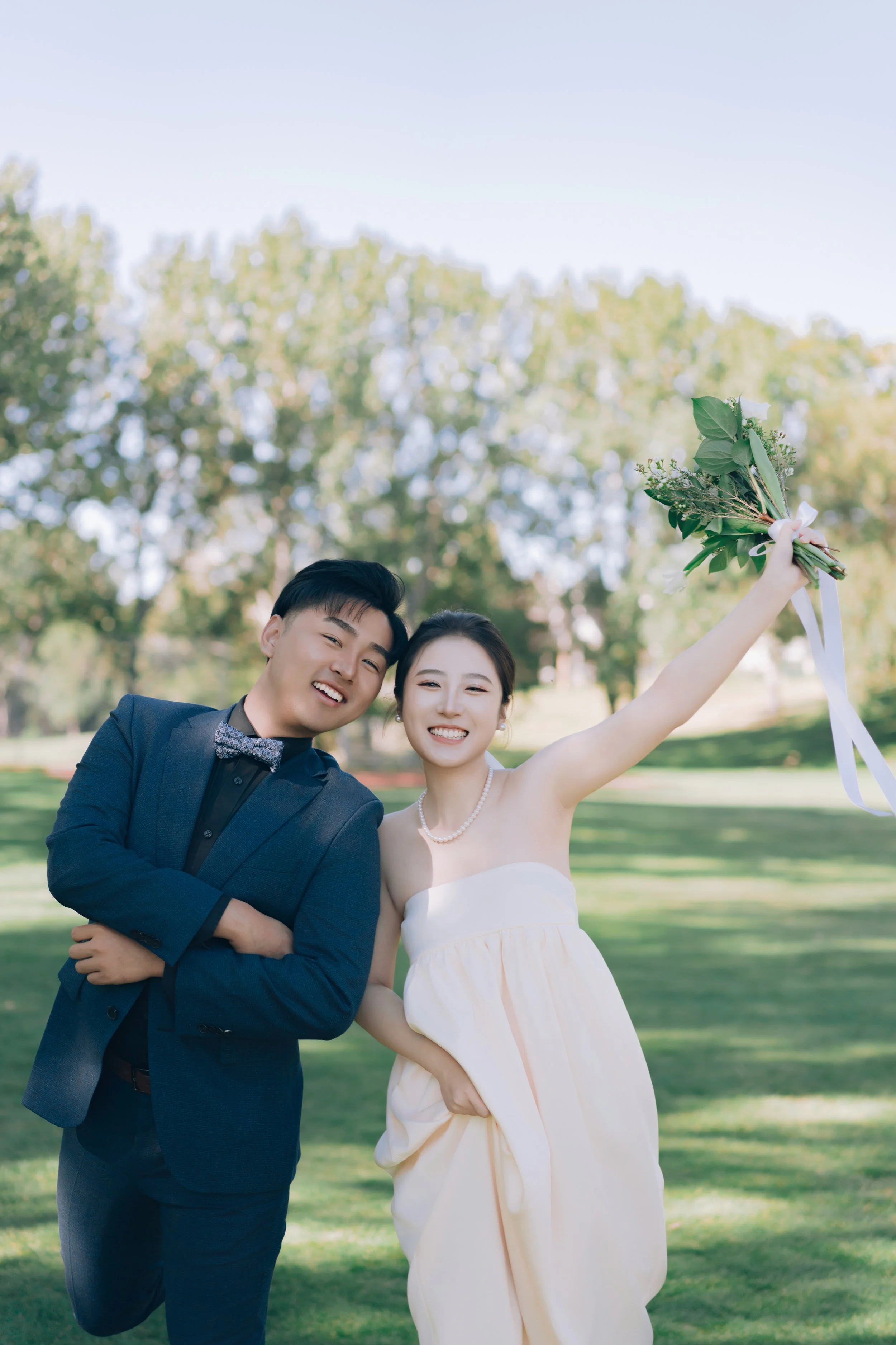 A young couple dressed in formal attire celebrating outdoors in a park with trees in the background. The woman is holding a bouquet of flowers raised in the air, smiling joyfully, wearing a strapless dress and pearl necklace. The man is wearing a dar