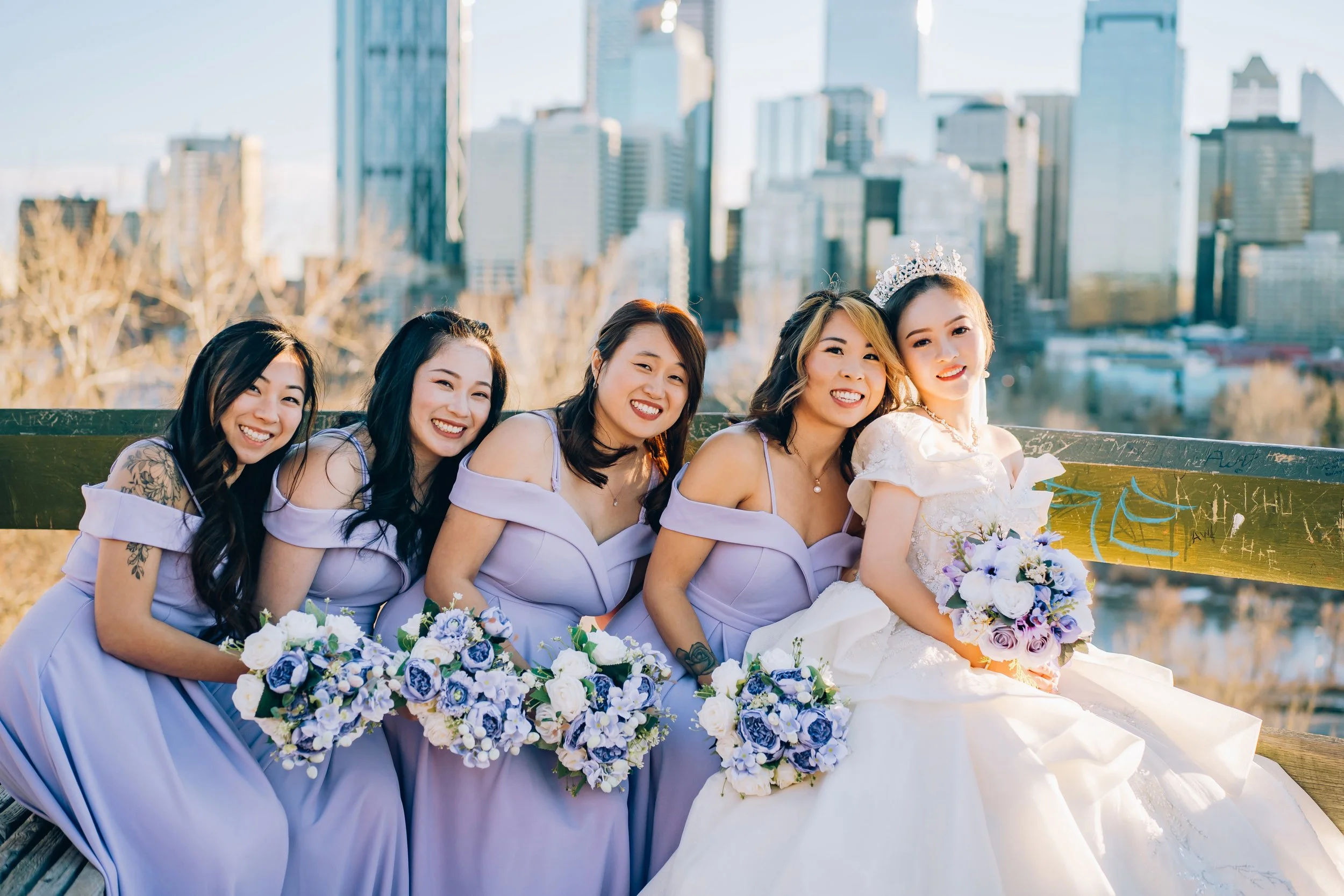 Bride in a white wedding dress holding a bouquet, surrounded by five bridesmaids in lavender dresses, all smiling and holding bouquets, with a city skyline in the background.
