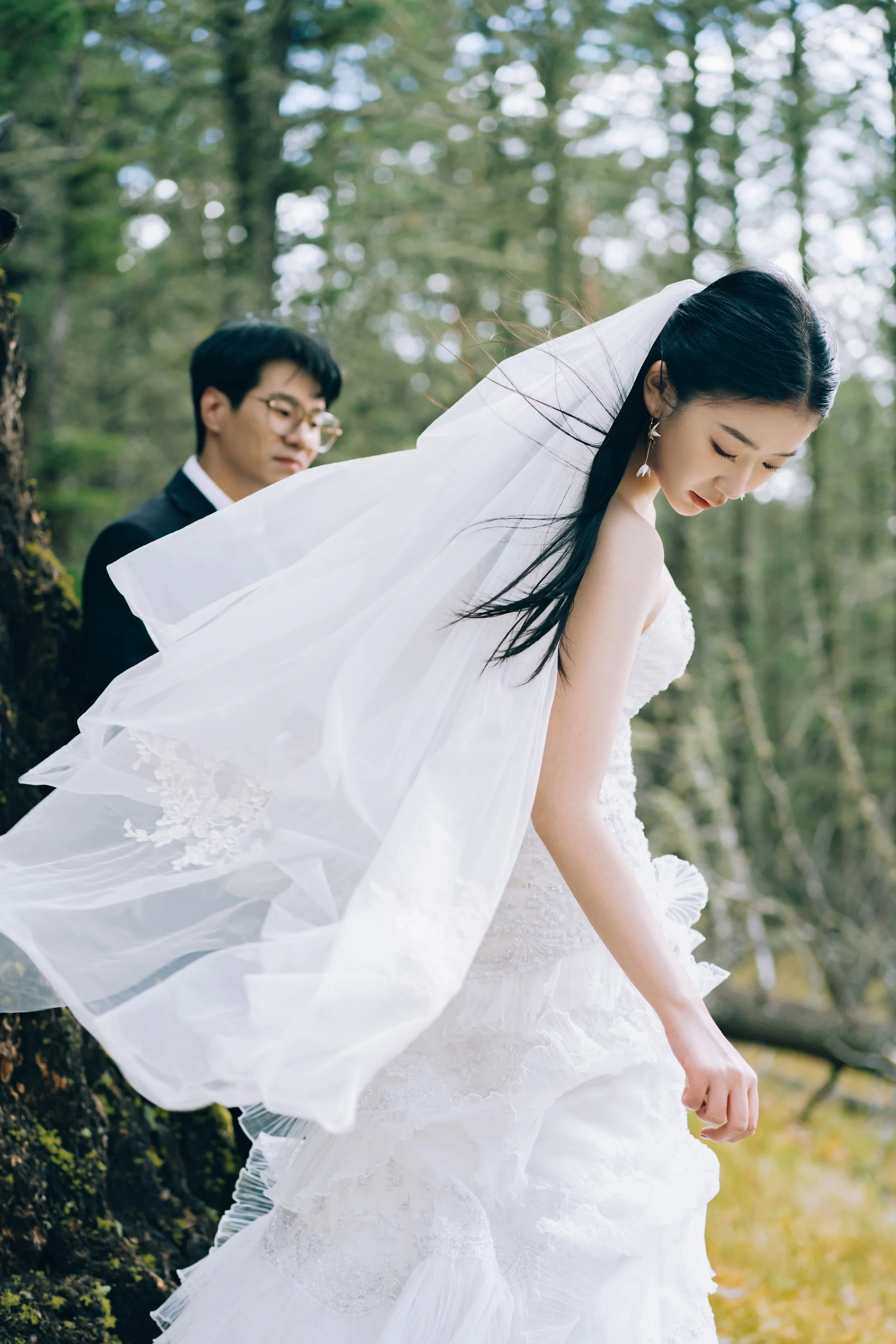 A bride in a white wedding dress and veil stands outdoors in a forest, with a groom in a dark suit and glasses in the background.