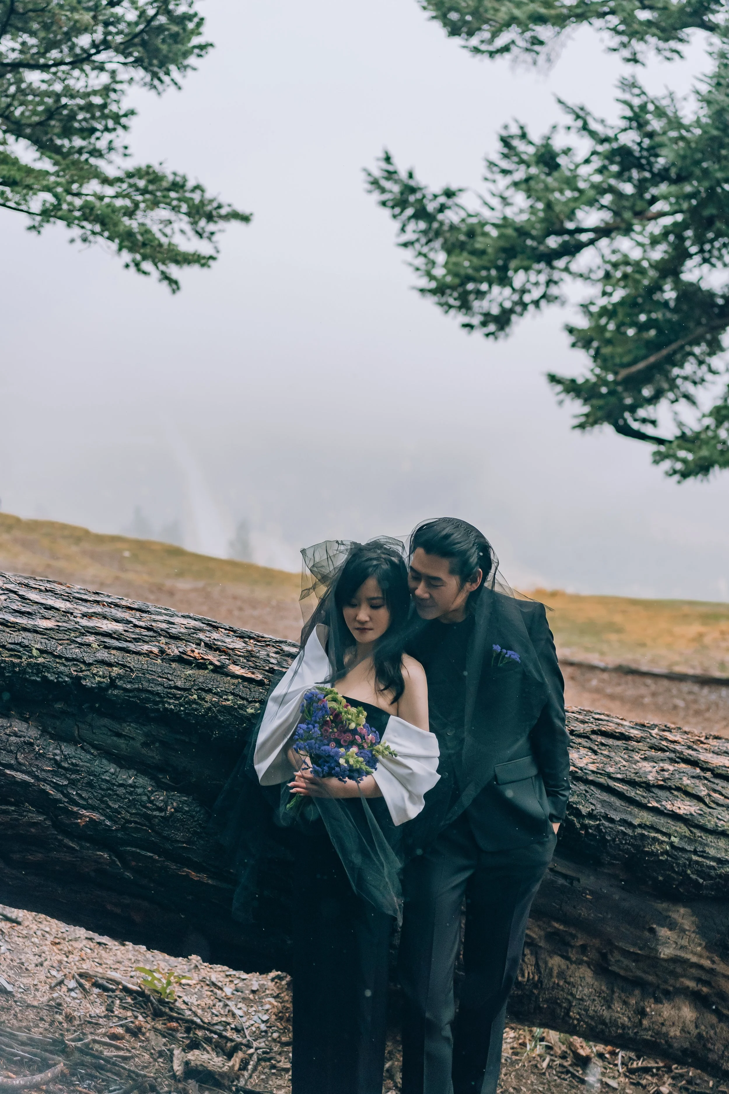 A couple dressed in formal attire, the woman holding a bouquet, standing beside a fallen tree in a forested area, with a misty background and trees overhead.