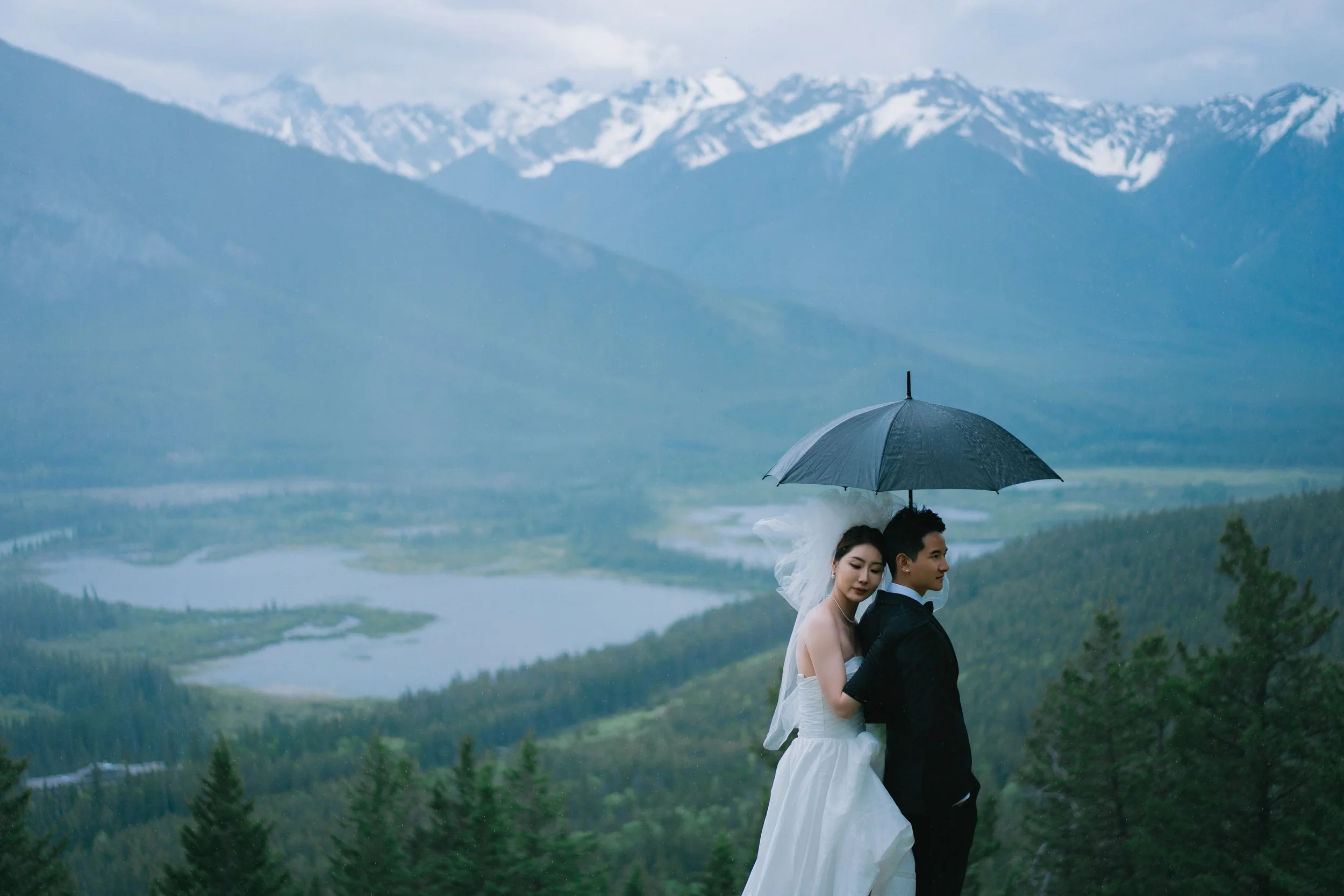 A bride and groom standing in the rain with an umbrella against a mountainous landscape, snow-capped peaks, forested hills, and a lake in the distance.