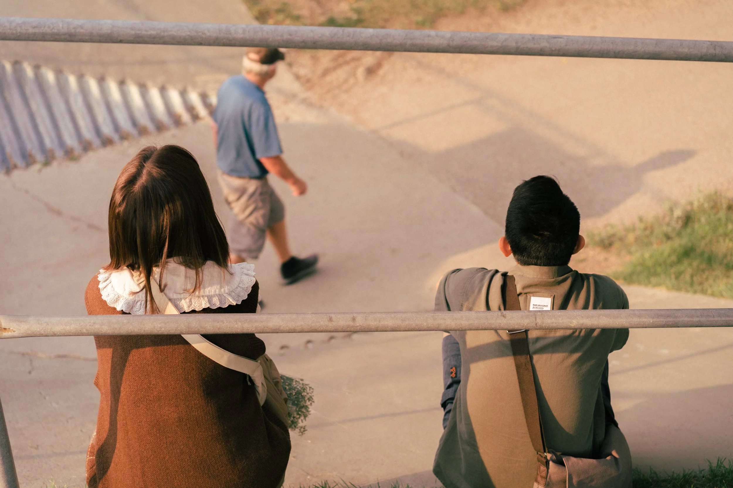 Two children are sitting on a ledge, watching an older man and young boy walking on the sidewalk in a park or schoolyard. The girl has shoulder-length dark hair and wears a brown sweater with a white lace collar, while the boy has short dark hair and