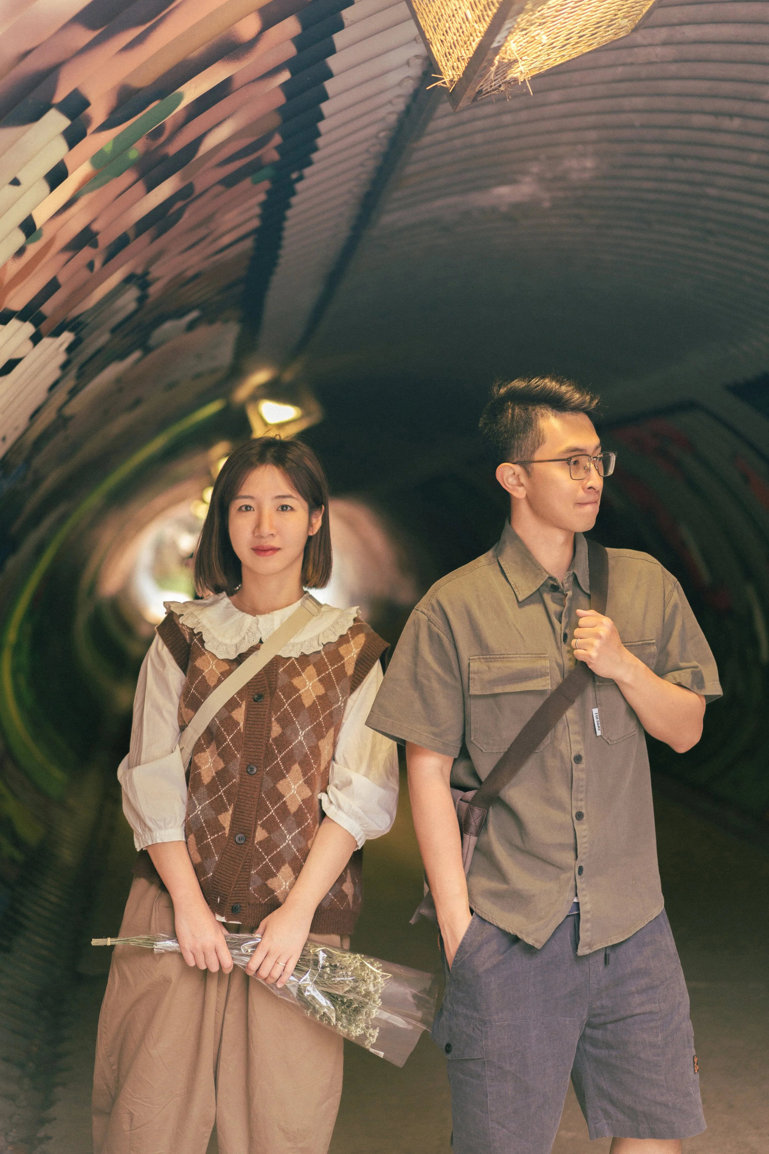 A young woman holding a bouquet of flowers and a young man wearing glasses walk through a tunnel with colorful reflective walls.