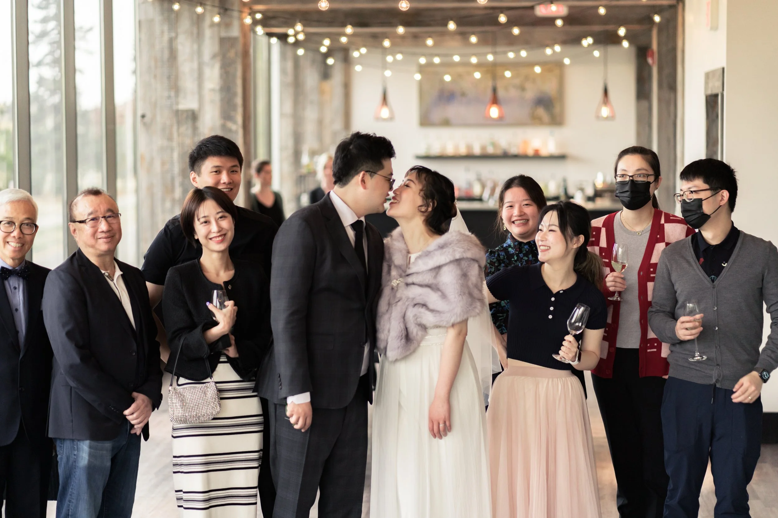 A diverse group of people at a wedding reception, with a bride and groom kissing in the center, surrounded by guests holding glasses of wine, all smiling and celebrating indoors.