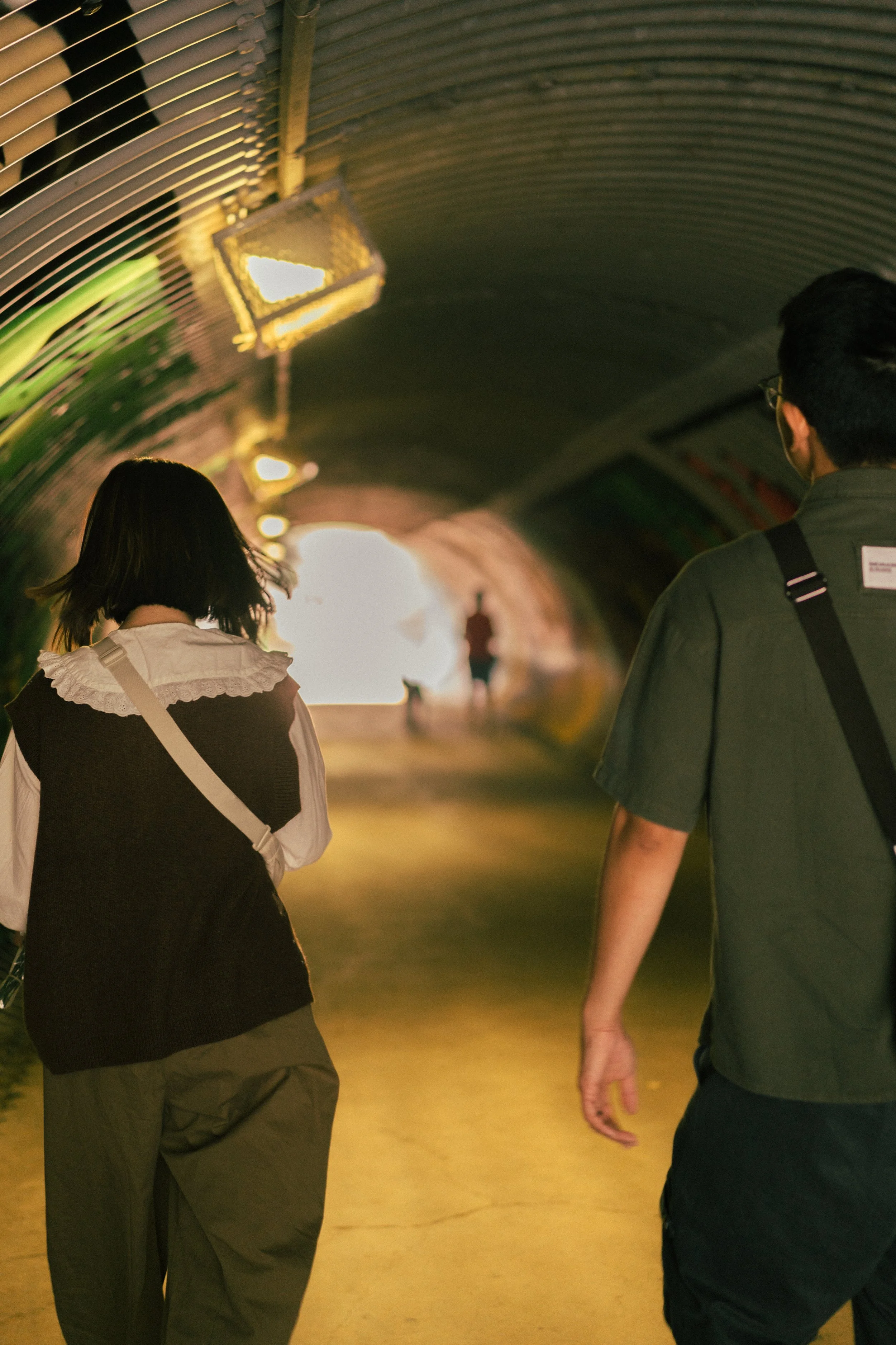 Two people walking through a tunnel with warm lighting, heading toward the bright exit or daylight outside.