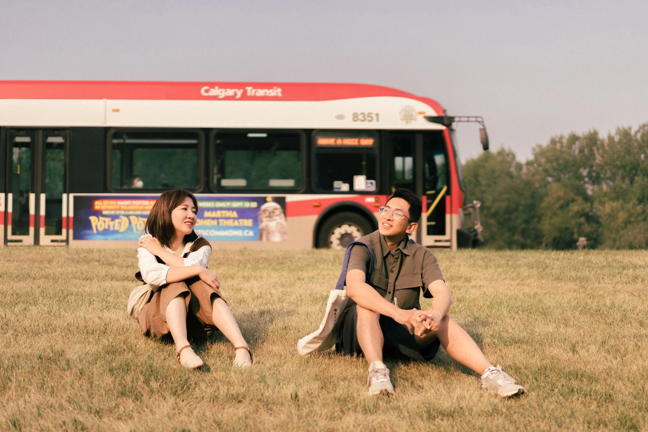 Two young people sitting on the grass near a red Calgary Transit bus, having a conversation on a sunny day.