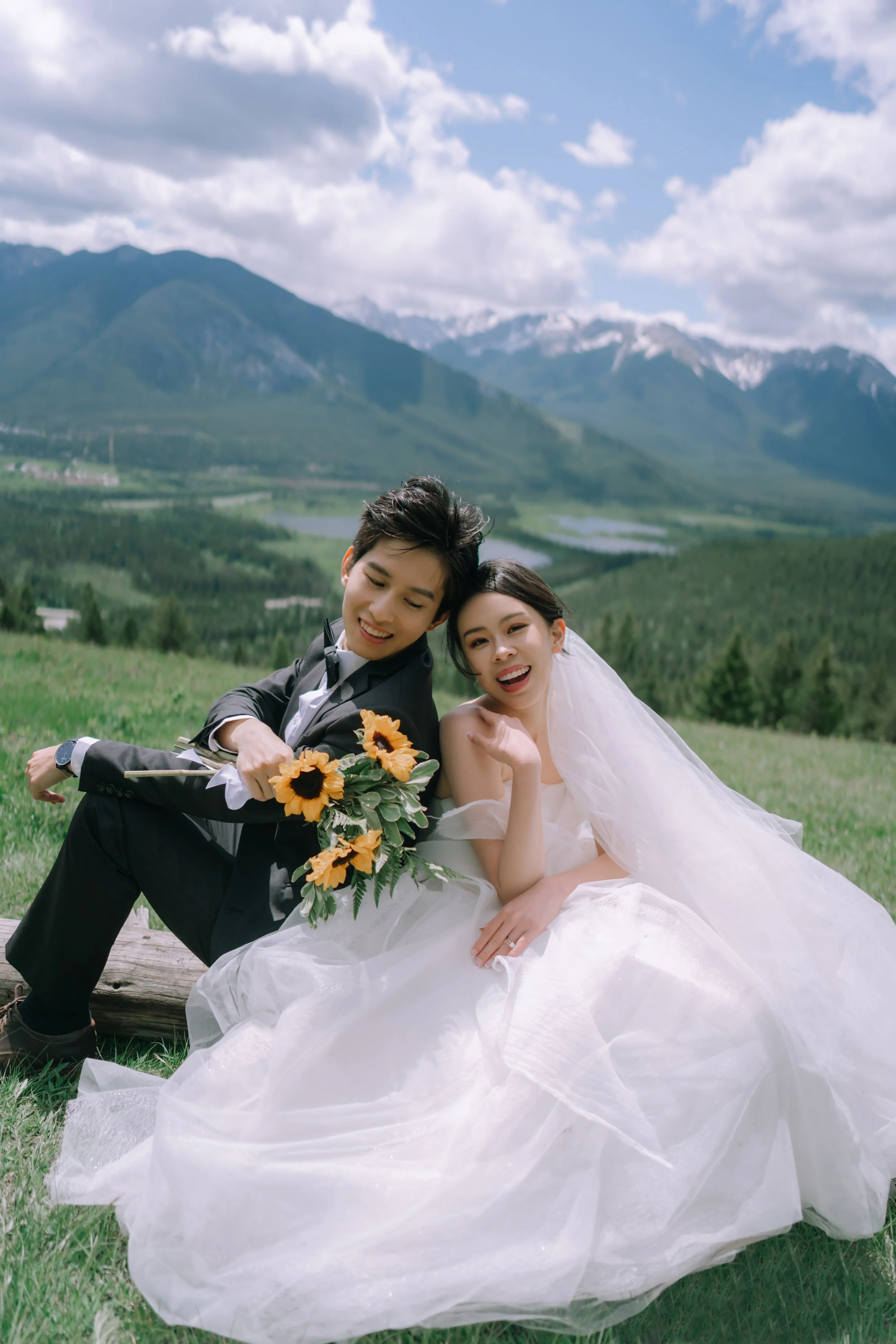 A smiling couple dressed in wedding attire sitting on a log in a lush green field with mountains, clouds, and blue sky in the background, the groom holding a bouquet of sunflowers.