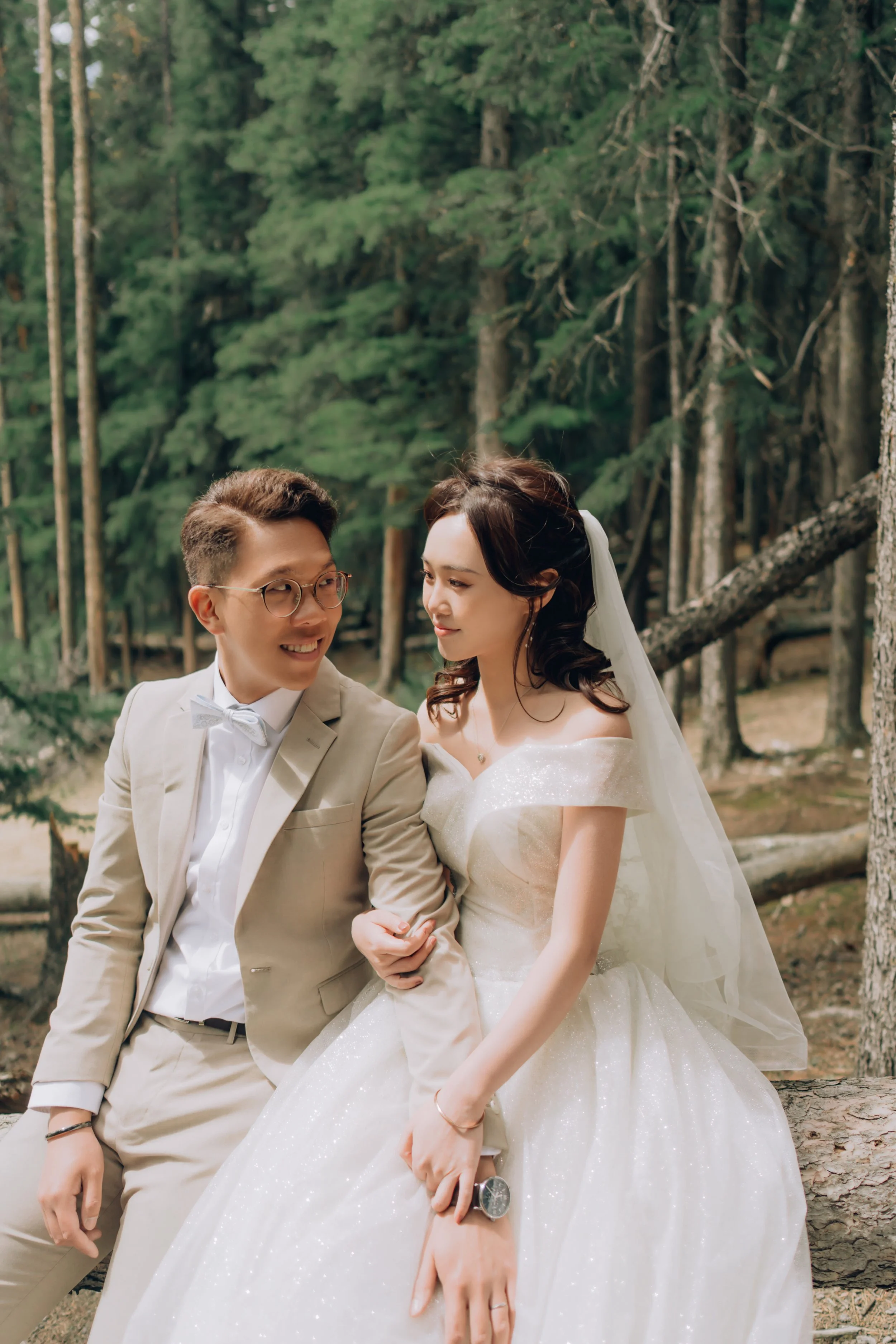 A bride and groom sitting outdoors among trees, smiling at each other, with the bride in a white wedding dress and the groom in a beige suit.